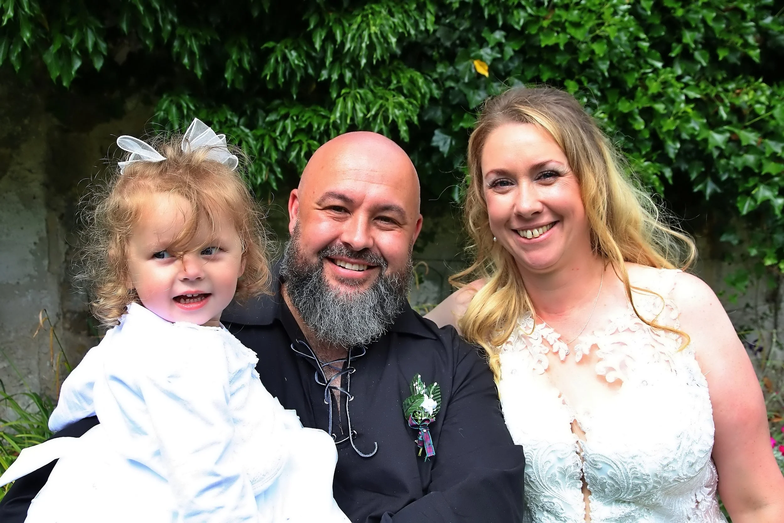 A family of three posing outdoors in front of green foliage. A young girl with curly hair and a white dress is on the left, a man with a beard and black shirt is in the middle, and a woman with long blonde hair and a white lace dress is on the right.