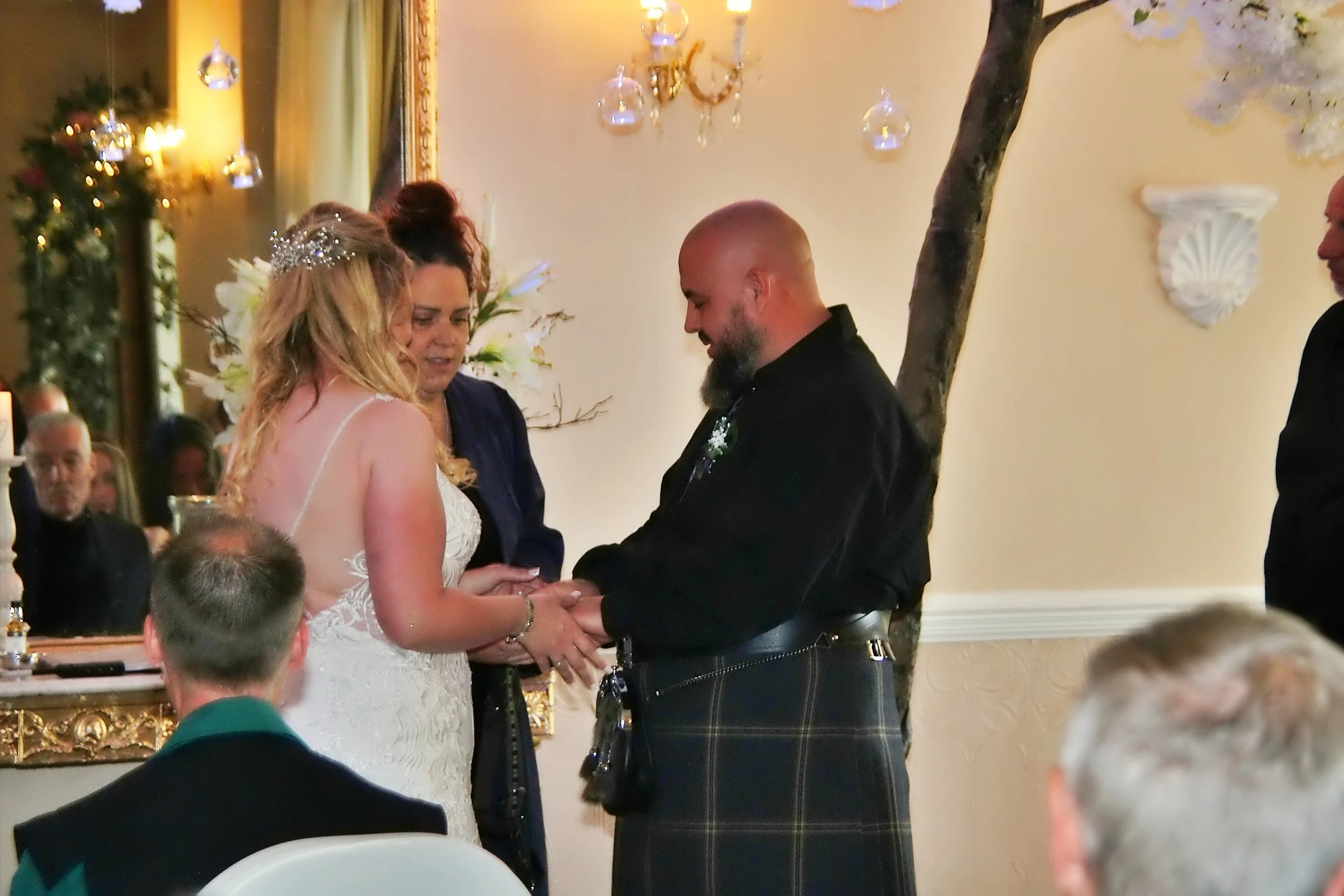 A couple exchanging wedding vows during a wedding ceremony, with an officiant and guests present. The bride is wearing a white lace wedding dress with a tiara, and the groom is in a black shirt and traditional tartan kilt. The setting has elegant dec