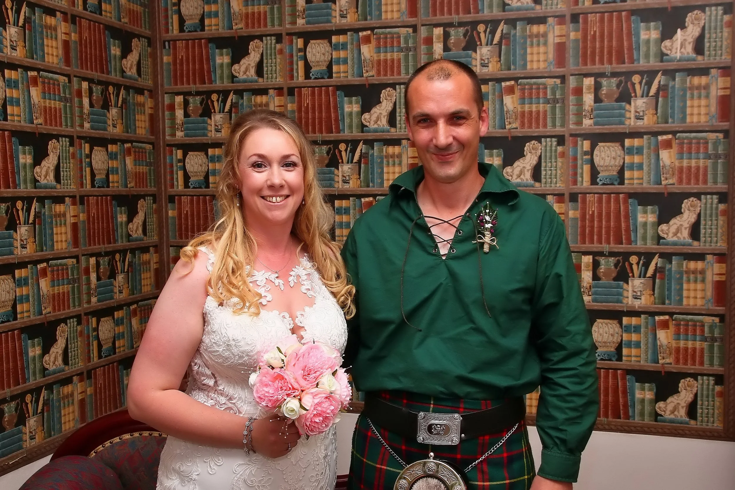 A smiling woman in a white lace wedding dress holding a pink and white bouquet, standing next to a man in traditional Scottish attire with a green shirt, tartan kilt, and decorative brooch, in front of a backdrop resembling a library with books and l
