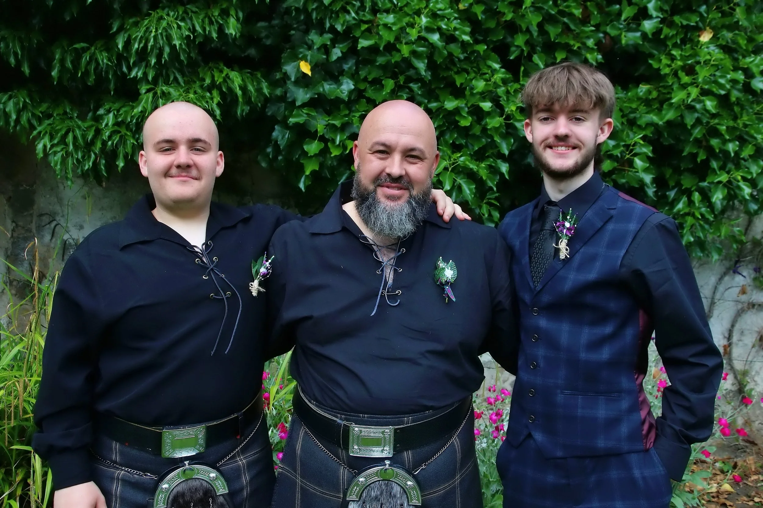 Three men dressed in traditional Scottish attire, standing outdoors with a green leafy background, smiling at the camera.