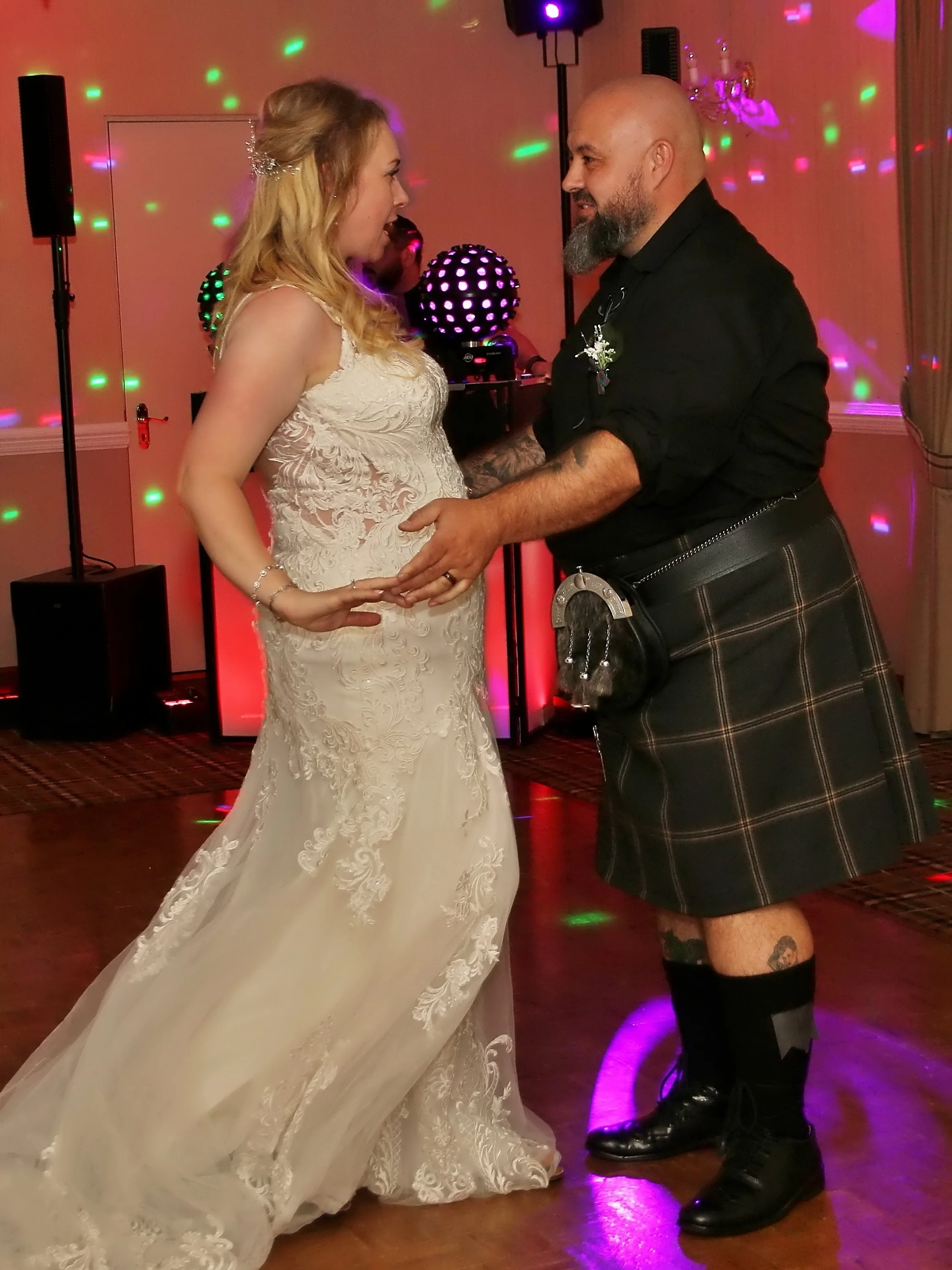 A bride and a man, possibly the groom, are dancing at a wedding reception with colorful lights and a DJ setup in the background.