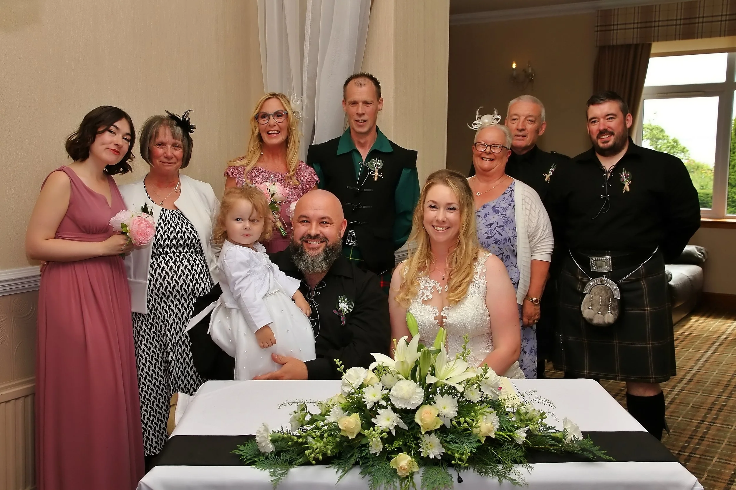 Group of people, including a bride and groom, celebrating at a wedding reception with floral arrangements on a table in front of them.