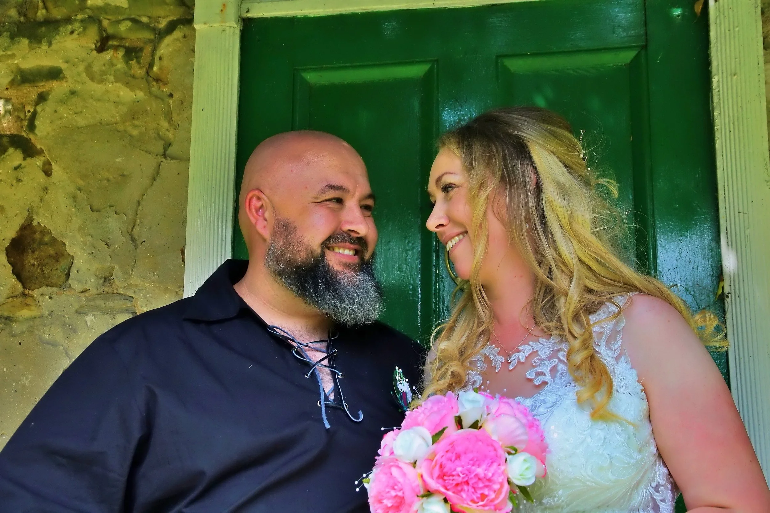 A man and woman in wedding attire smiling at each other, standing in front of a green door and stone wall, woman holding a bouquet of pink and white flowers.