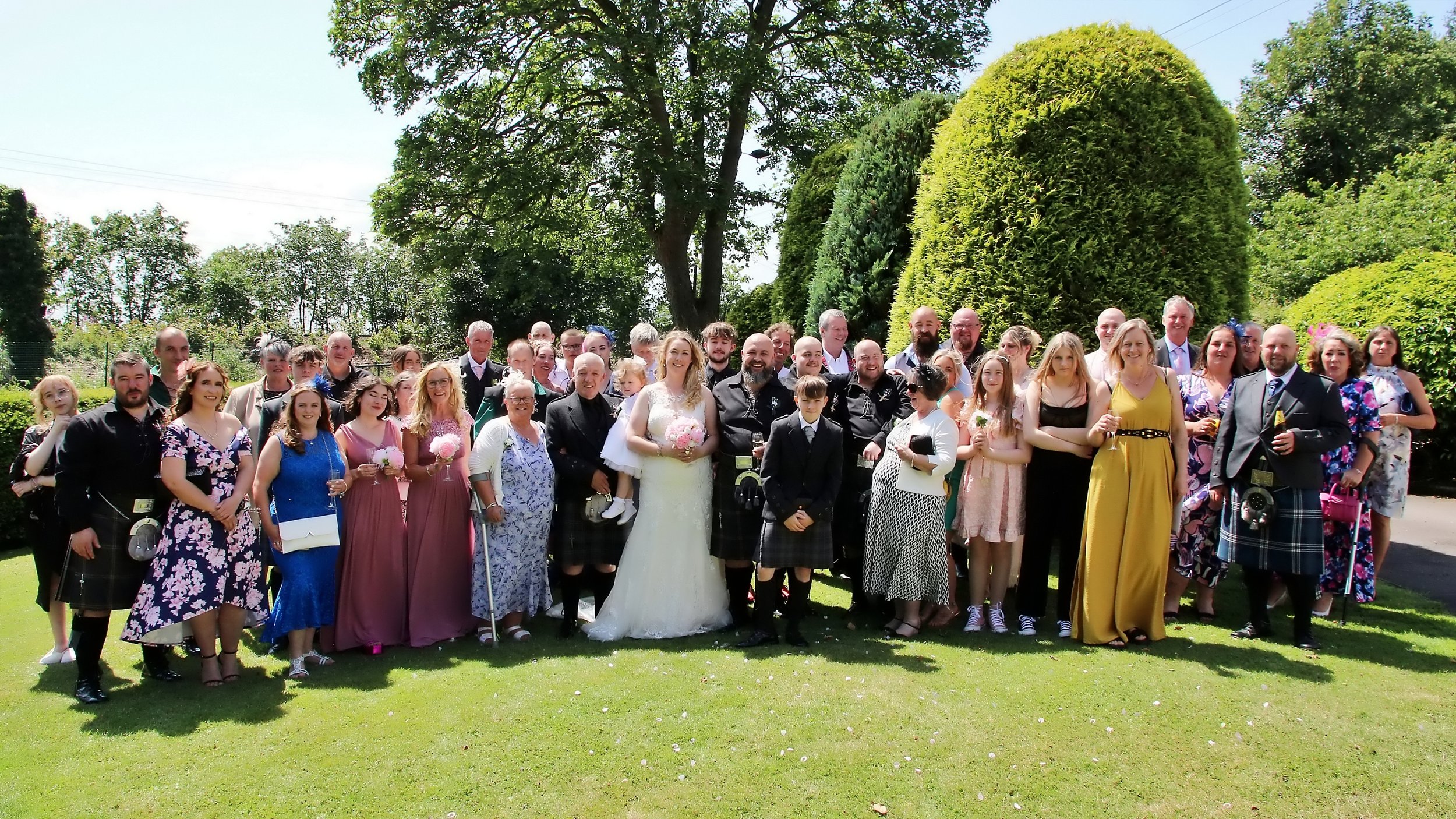A large group of people in colorful formal and traditional attire gathered outdoors on a sunny day, with green trees and bushes in the background, for a wedding celebration.