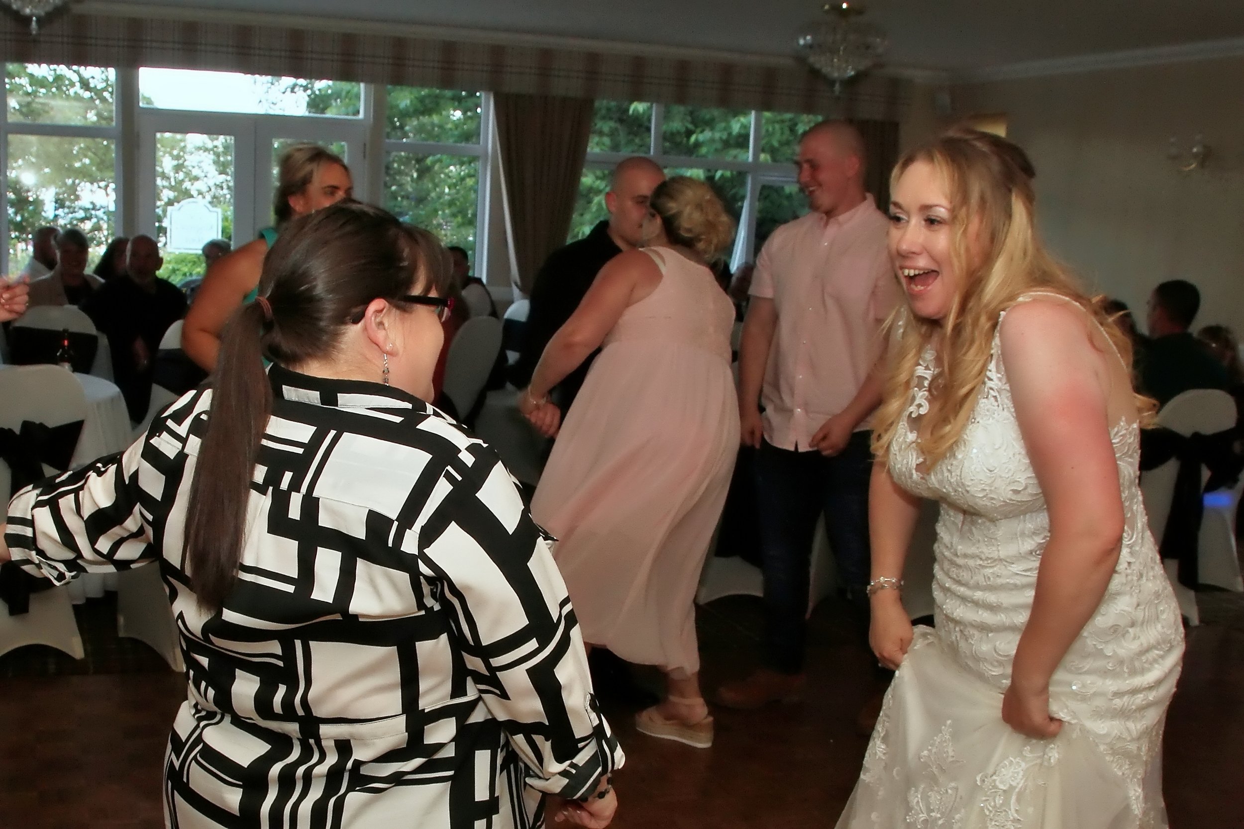 People dancing and socializing at a wedding reception or celebration in a decorated indoor venue with windows and trees outside.