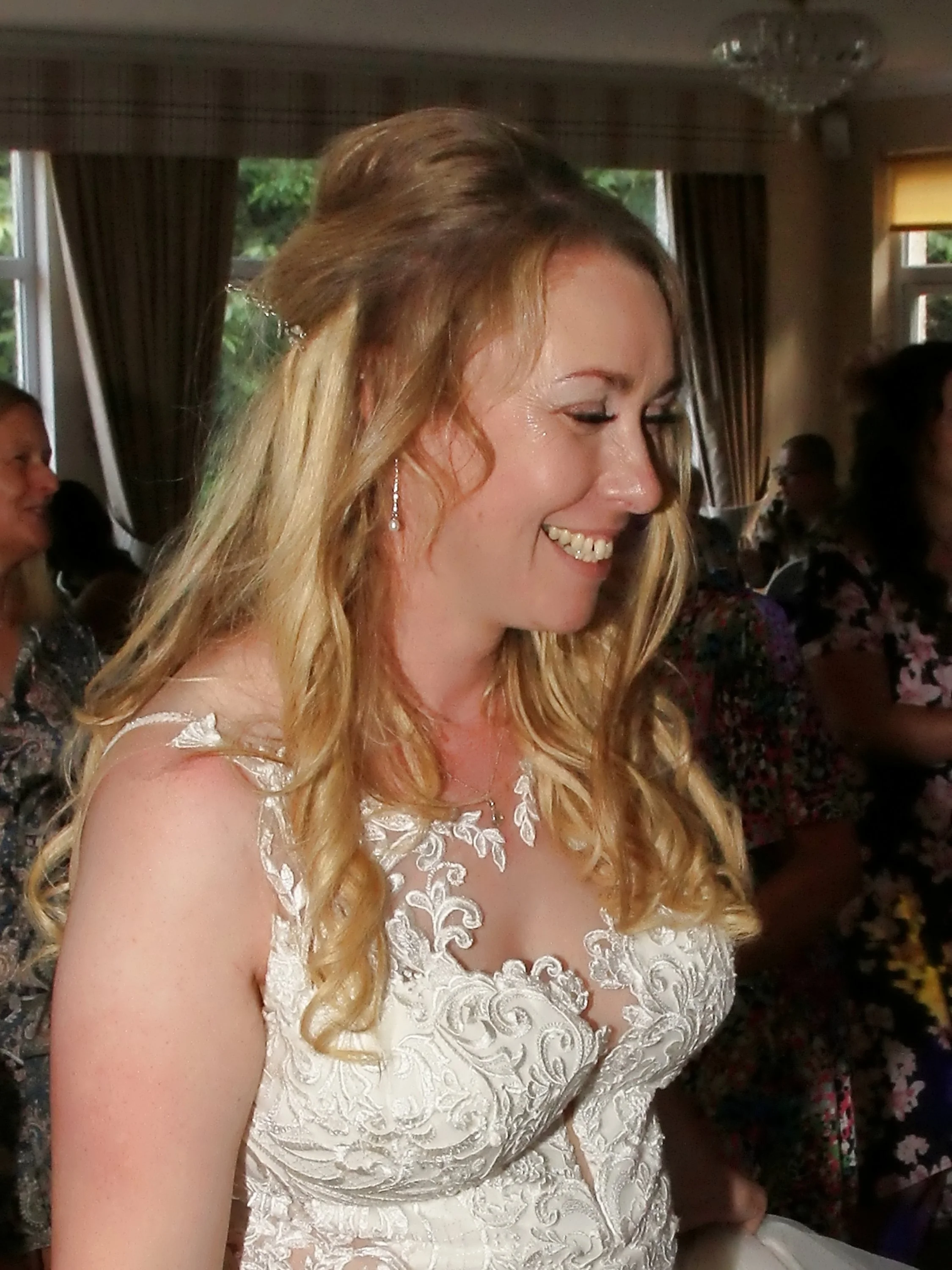 A woman with blonde wavy hair smiling, wearing a white lace dress with floral embroidery, at a social event indoors with other people in the background.