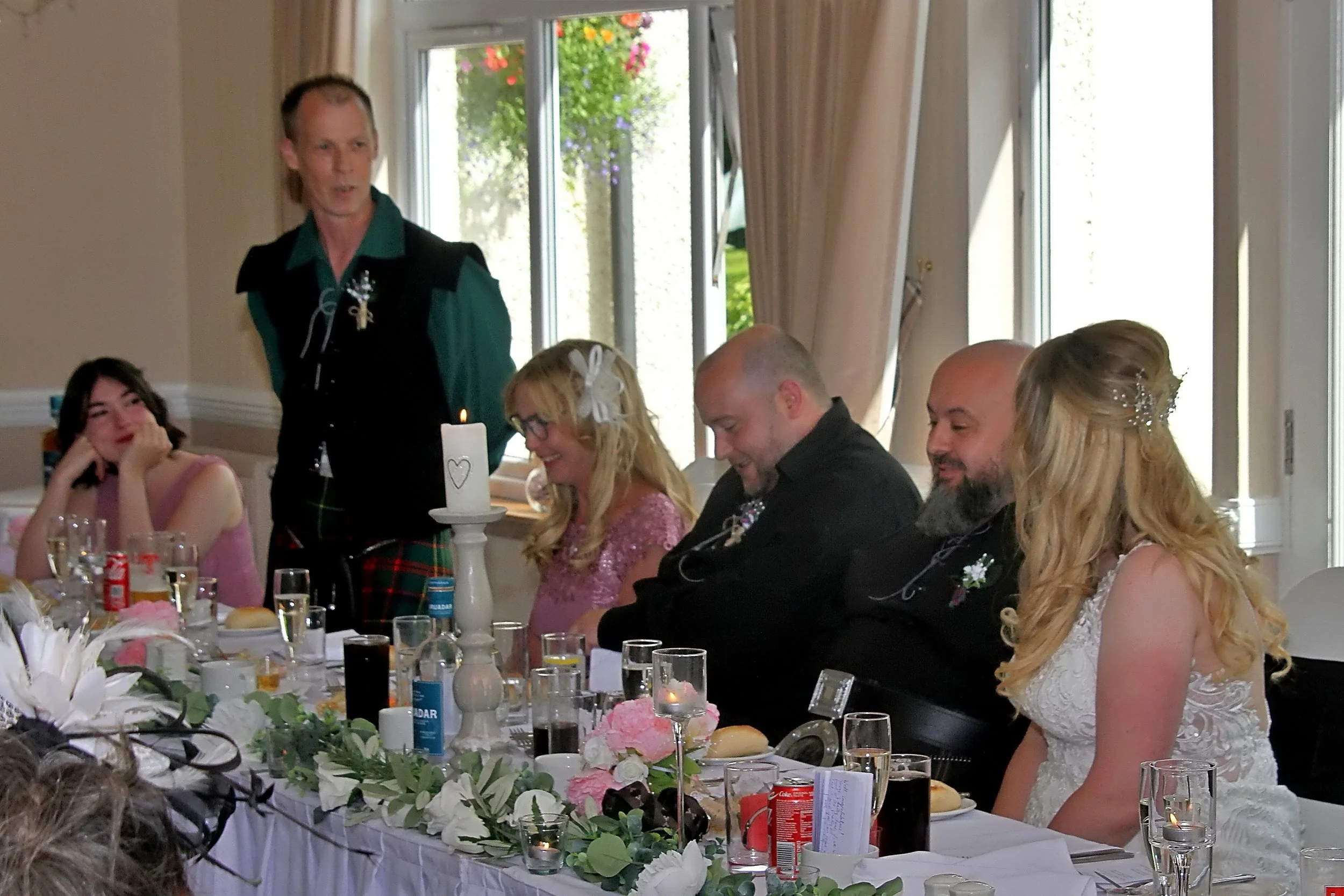 People sitting at a long table during a wedding reception, with a man in traditional Scottish attire standing and giving a speech.
