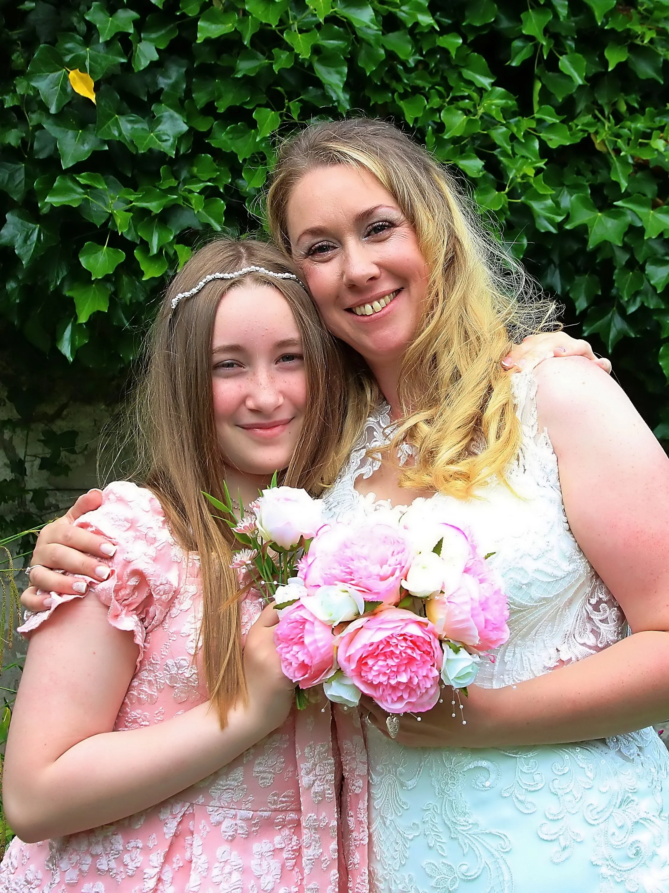 Two women, one older and one younger, dressed in elegant clothing, standing close together outdoors with a background of green ivy. They are smiling, and the younger woman is holding a bouquet of pink and white flowers.