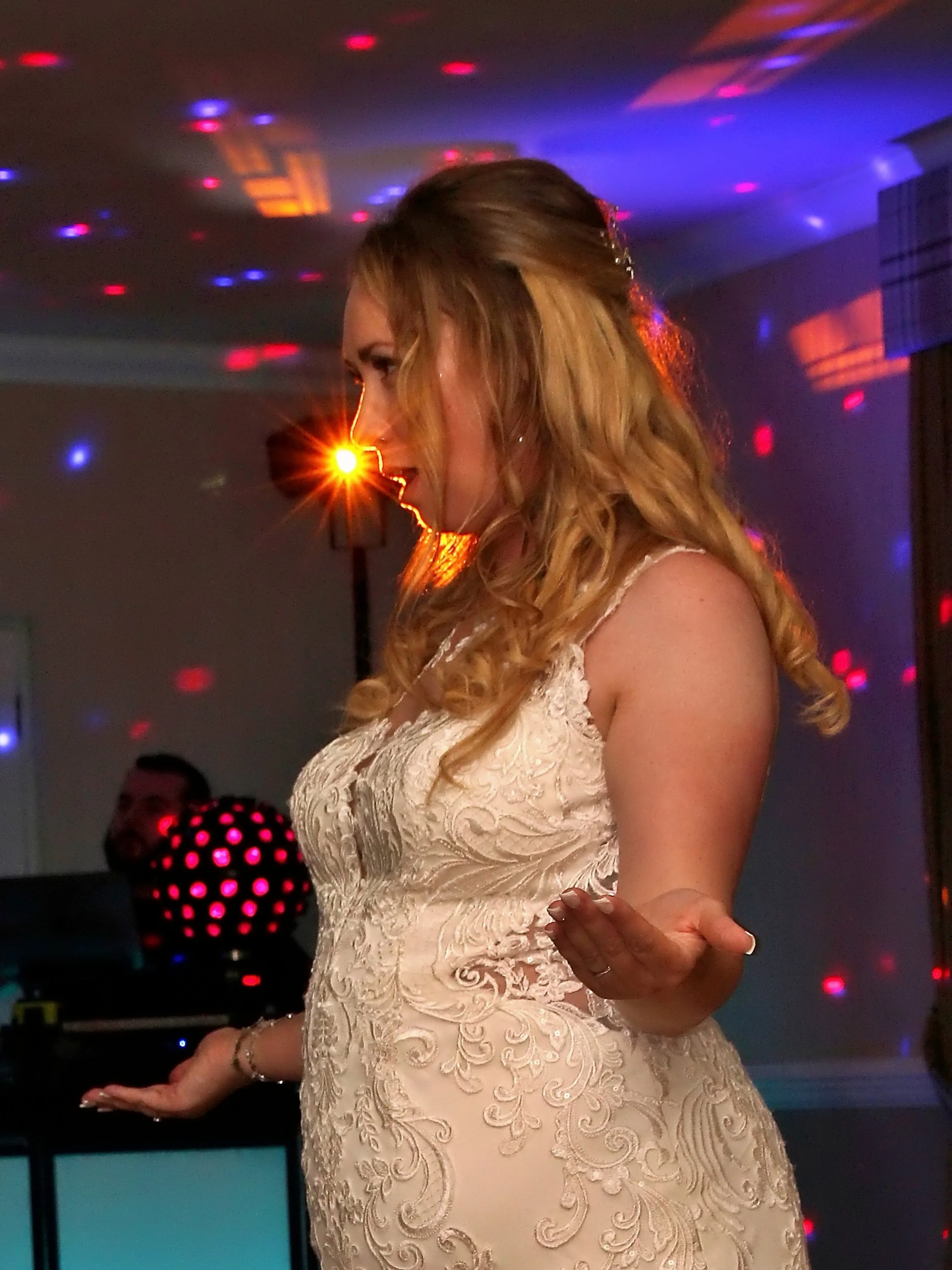 A woman with long, curly blonde hair dancing at a party or celebration. She is wearing a beige, embroidered dress and appears to be enjoying herself, with colorful party lights and a DJ in the background.