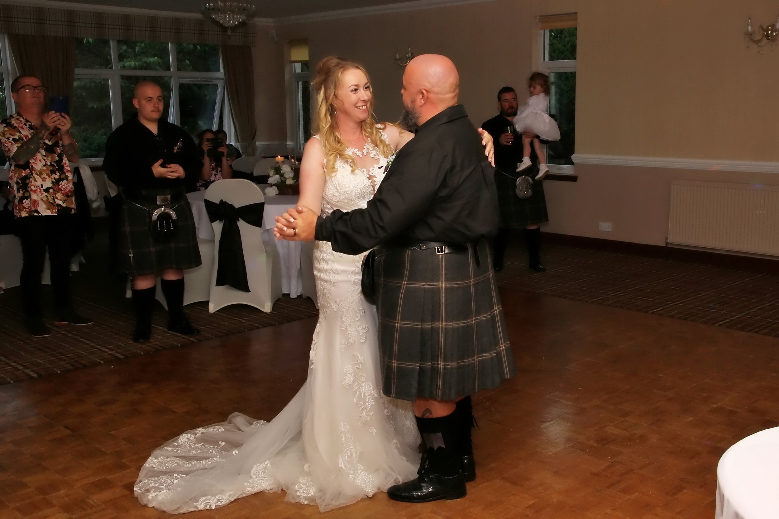 A bride and a groom dancing at their wedding reception in a decorated hall. The bride is wearing a white lace wedding gown, and the groom is in a black shirt and traditional Scottish kilt. Guests are watching and taking photos.