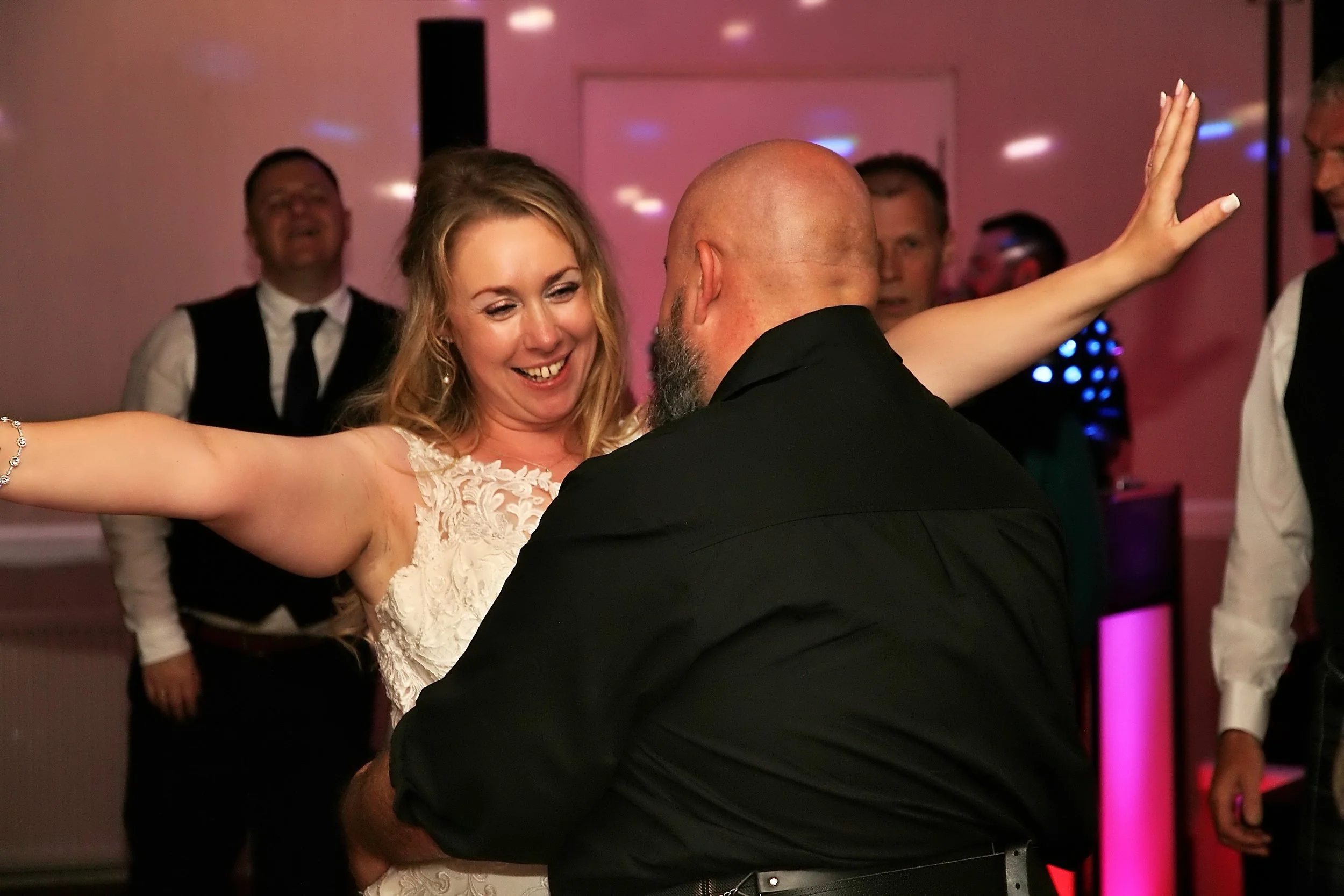 A joyful woman in a wedding dress dancing with a man at a celebration, with others dancing and enjoying in the background.