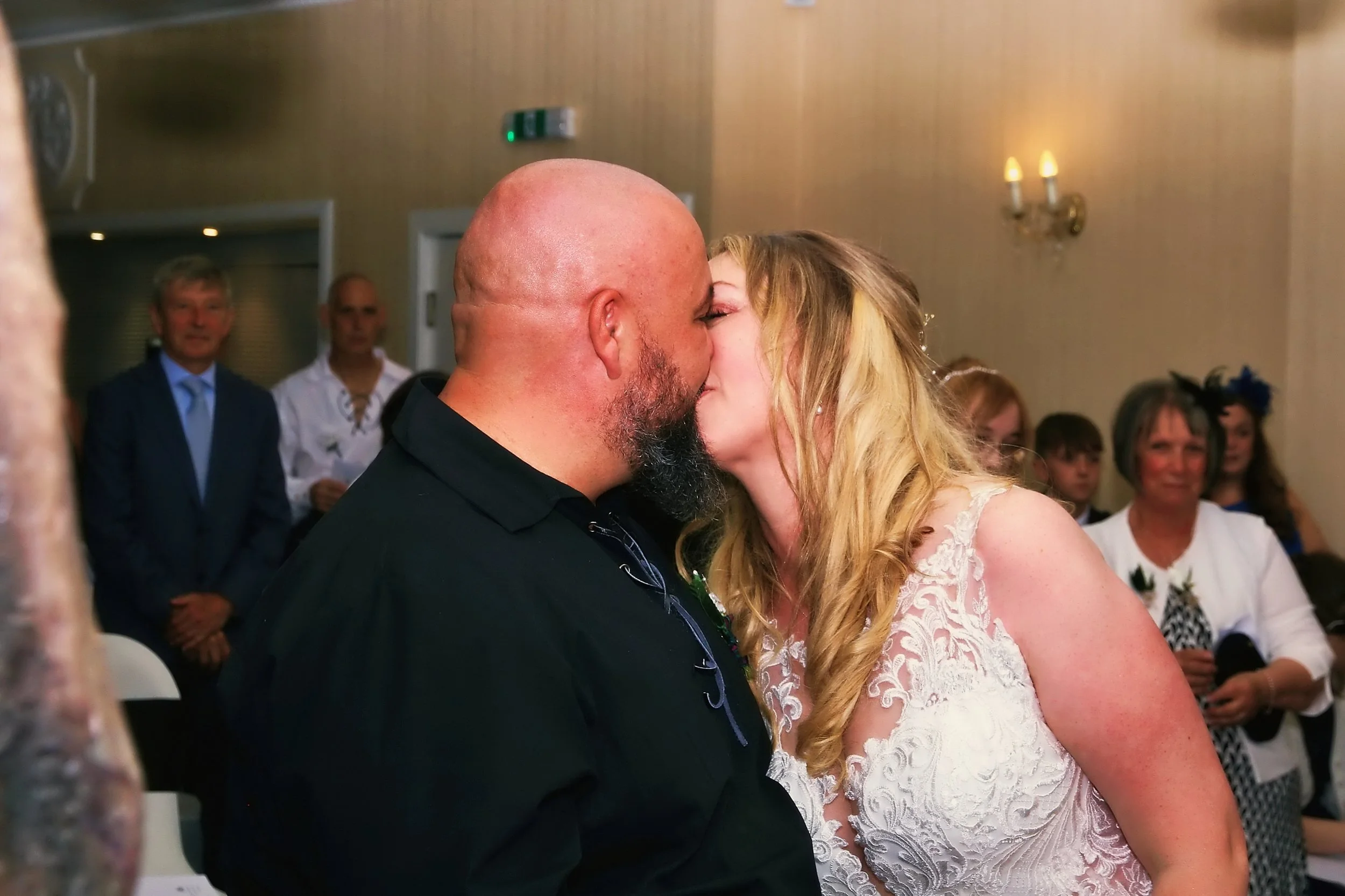 A couple kissing at a wedding reception, with guests watching in the background.
