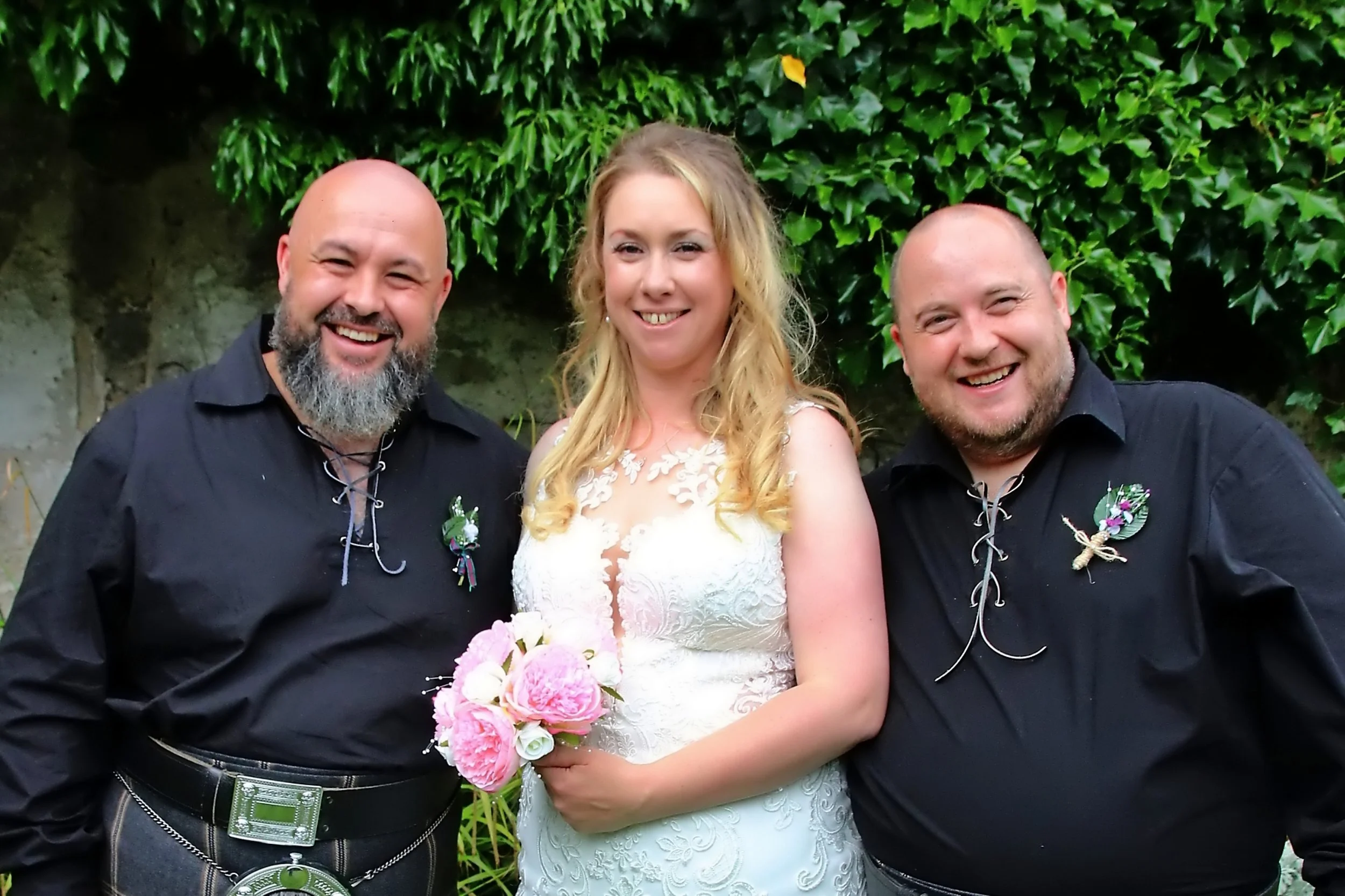 A bride with blonde hair holding a pink and white bouquet, standing between two men with bald heads and beards, all smiling outdoors in front of green foliage.