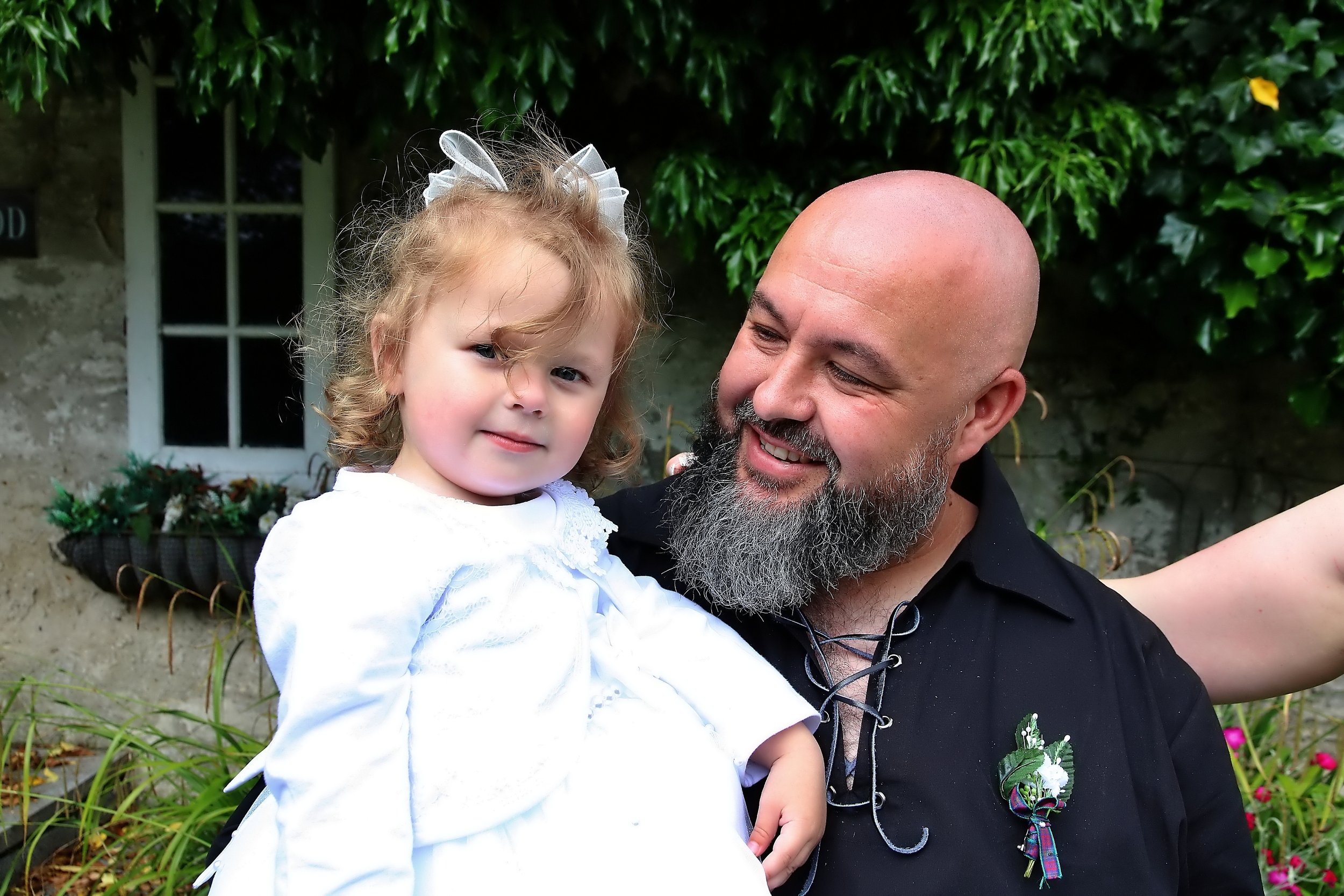 A man with a bald head and gray beard holding a young girl with curly hair and a white dress outdoors, with green foliage and a building in the background.