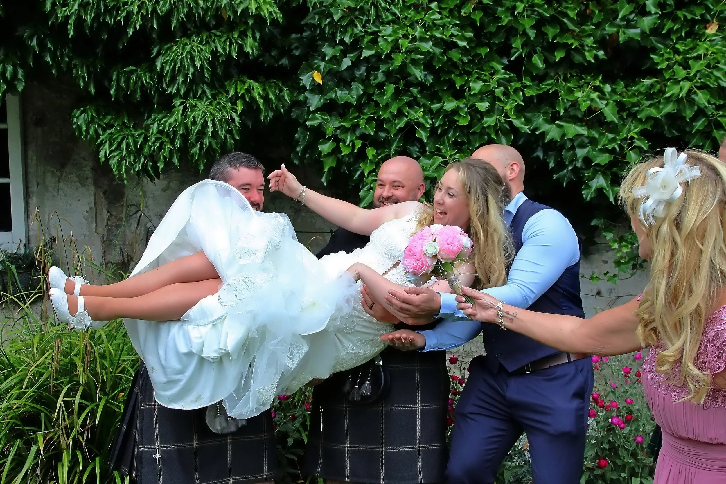 Group of wedding celebration people, with a woman in a wedding dress being held horizontally by men in kilts and surrounded by friends, outdoors with greenery in the background.