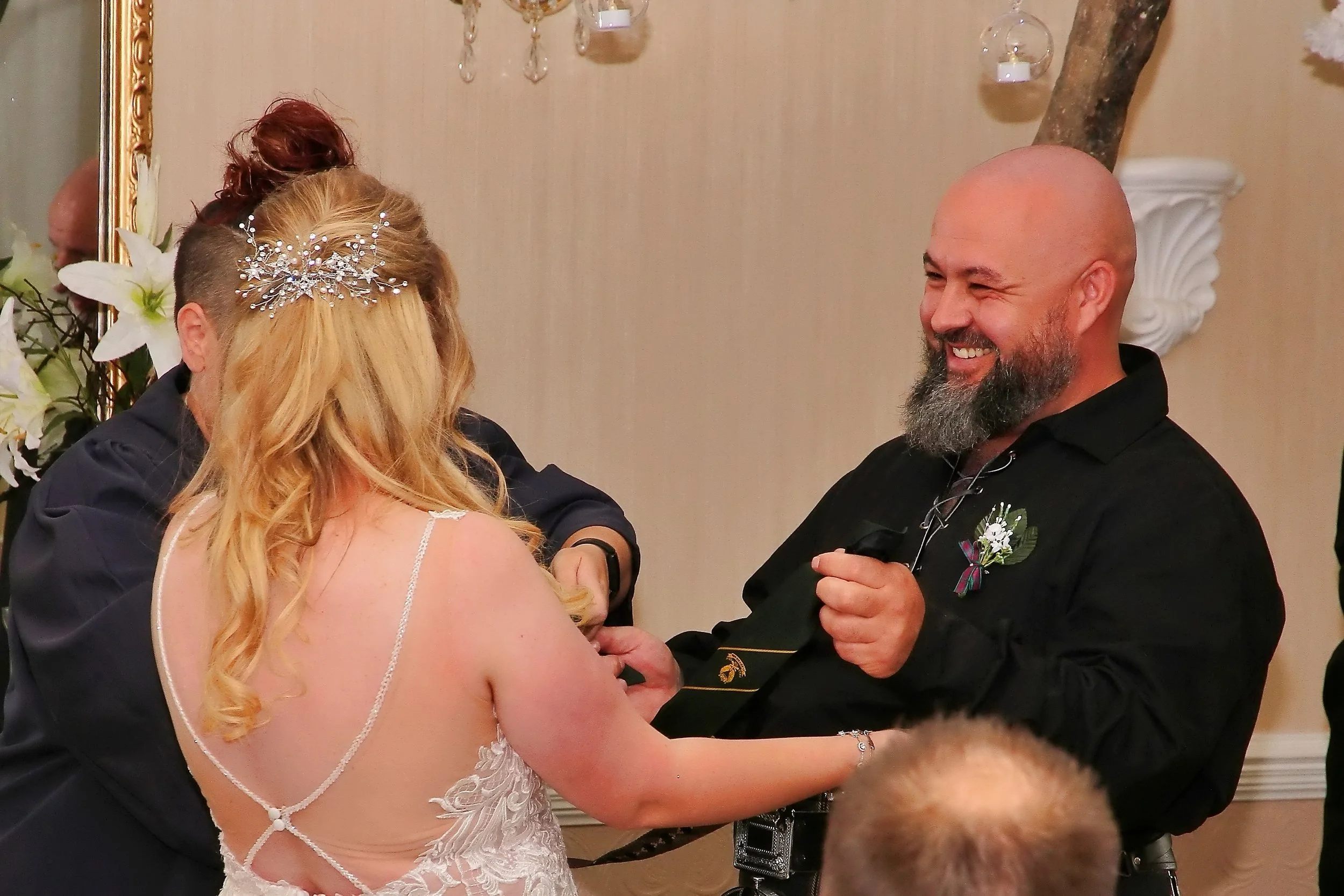 A bride and groom holding hands during their wedding ceremony, smiling at each other, with officiants present in an indoor setting decorated with flowers and wall ornaments.