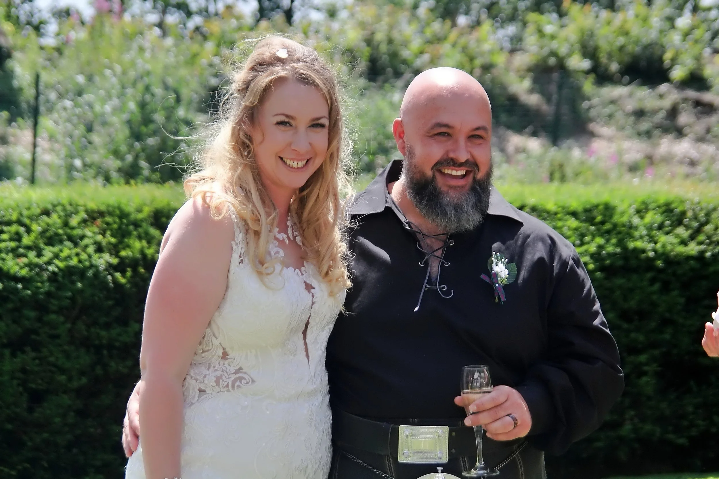 A smiling woman in a white wedding dress and a man with a bald head and gray beard in a black shirt, holding a glass of champagne, standing outdoors with green bushes and trees in the background.