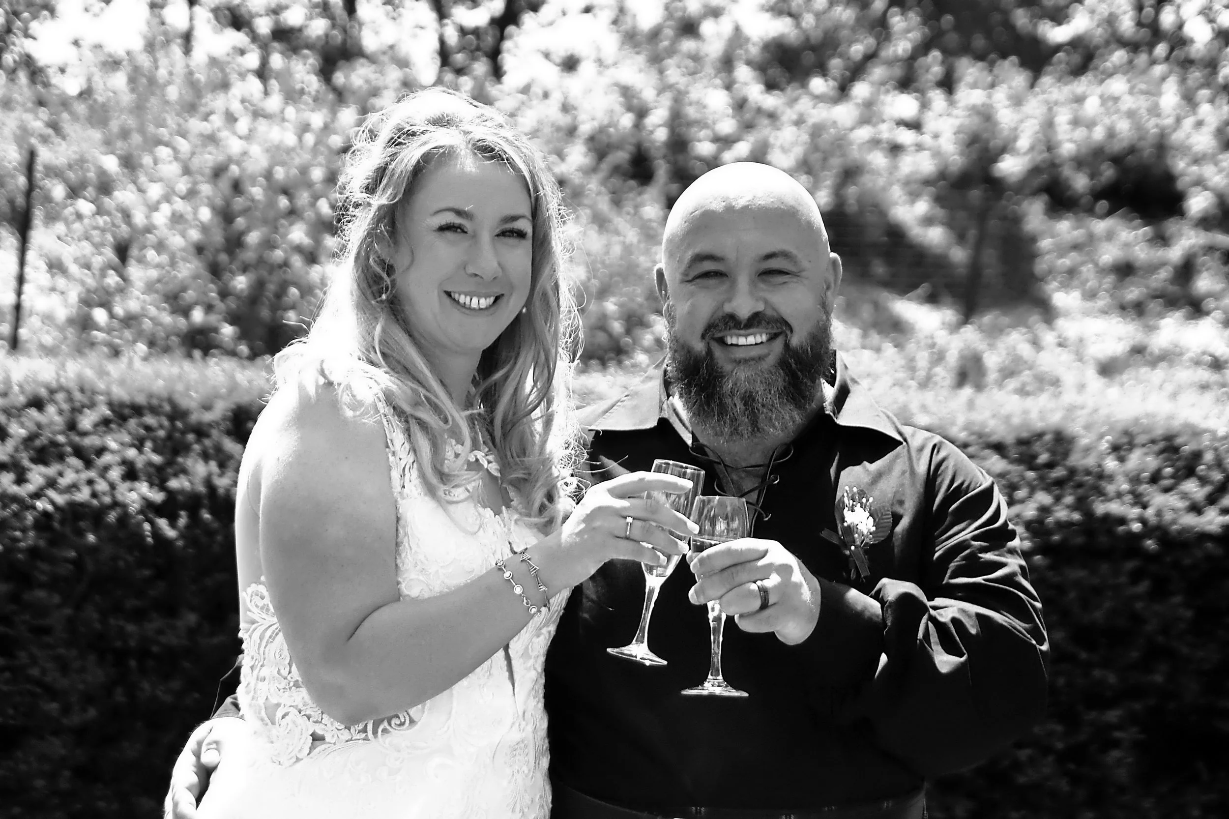 A smiling woman in a wedding dress and a smiling, bald man with a beard in a dark shirt are celebrating with champagne outdoors, with trees and foliage in the background.