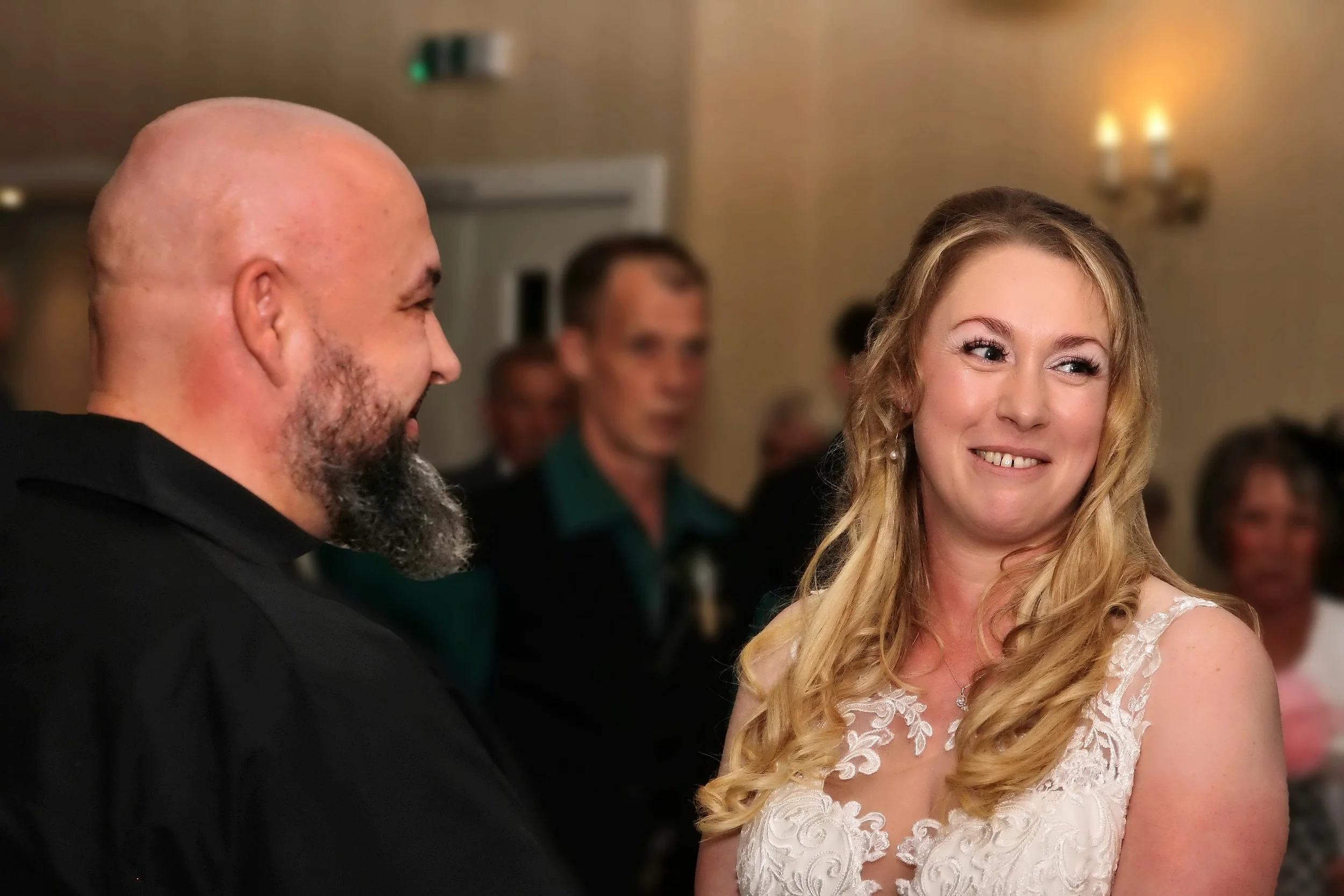 A man and woman smiling and looking at each other at a social event, with other people in the background.