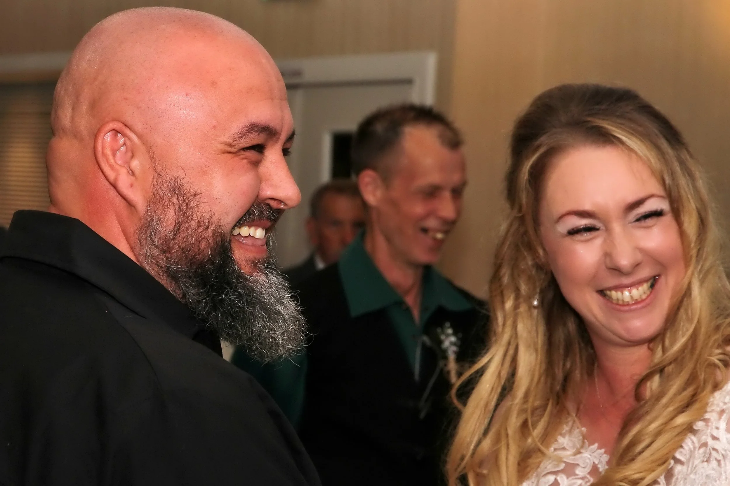 A group of four people smiling and laughing indoors, with focus on a bald man with a beard and a woman with long blonde hair in the foreground.