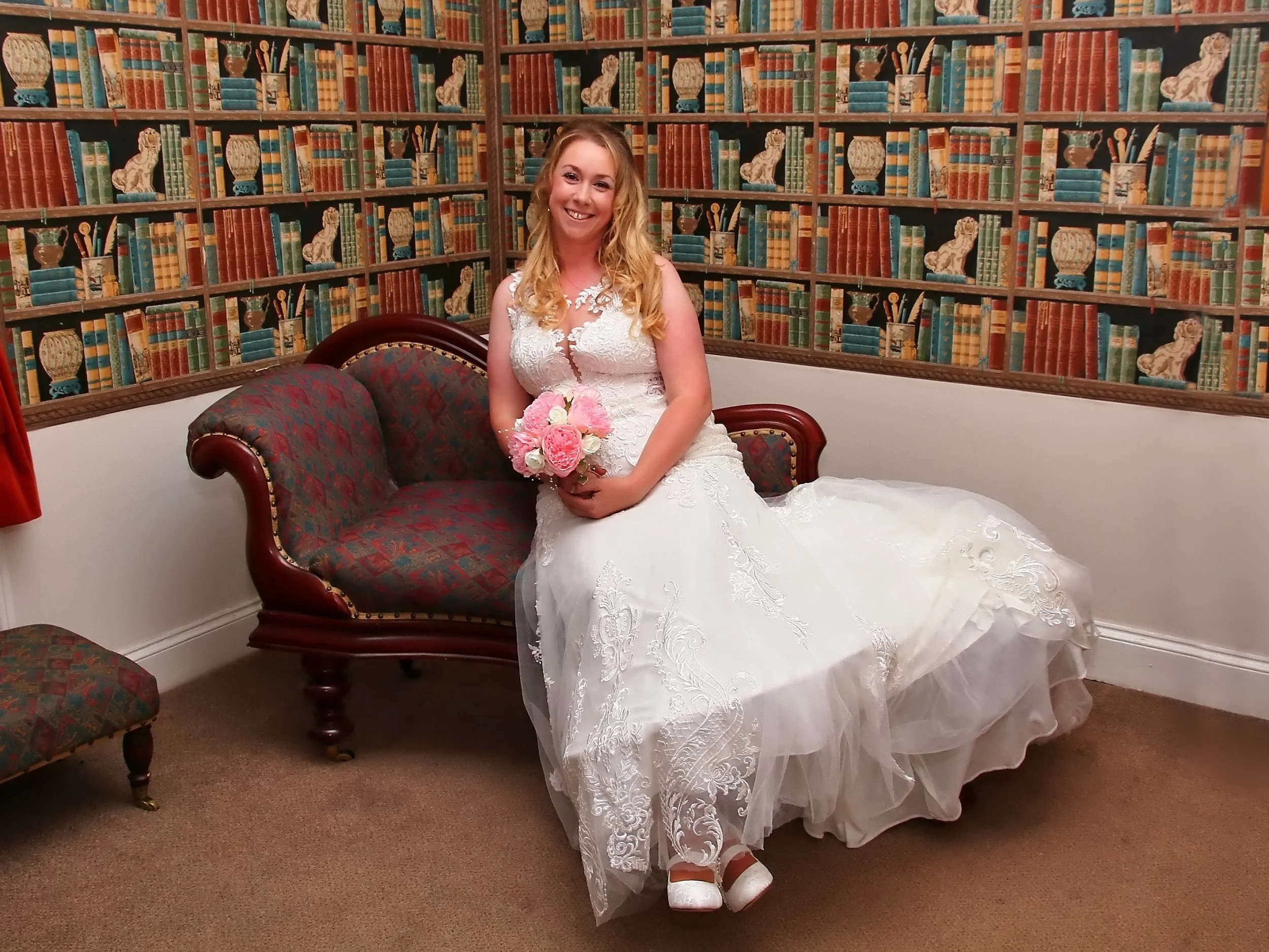 A woman in a wedding dress sitting on a vintage-style sofa, holding a pink and white bouquet, in front of a wallpaper with a bookshelf pattern with various books and decorative items.