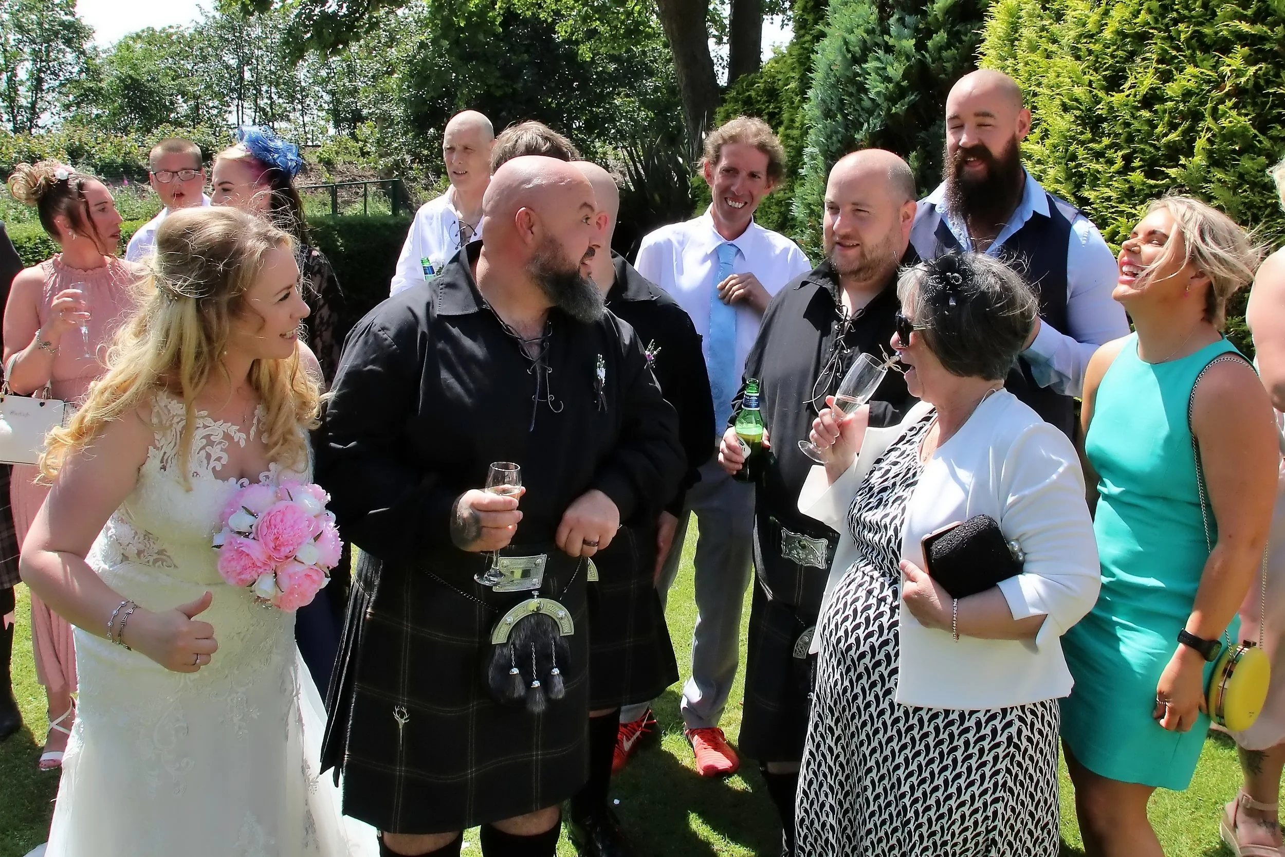 Group of people at an outdoor wedding celebration, with some holding drinks and engaging in conversation, surrounded by greenery and trees.