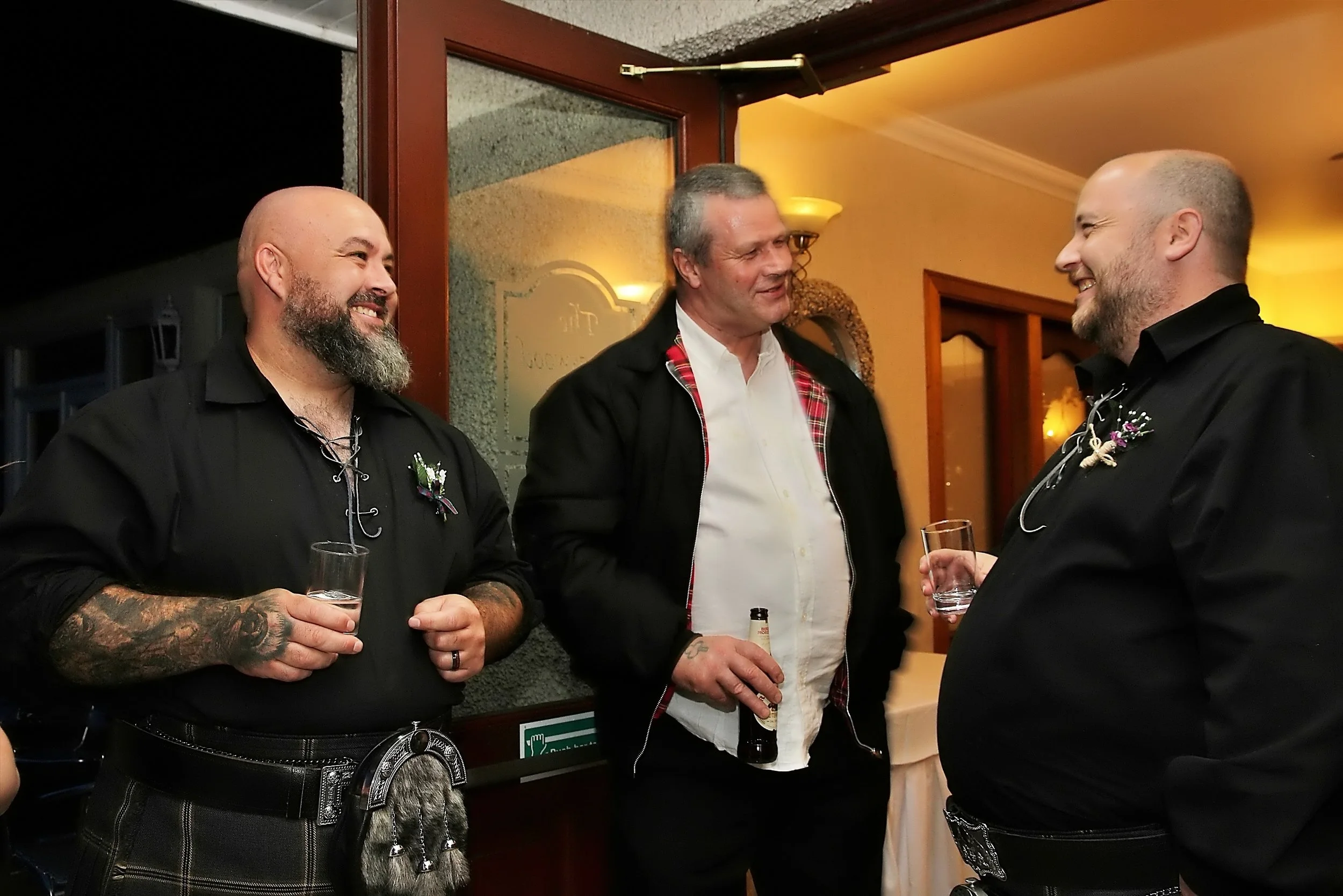 Three men engaged in conversation at an indoor social gathering, with two of them holding drinks. The man on the left has a bald head and beard, with tattoos on his arm, wearing a black shirt and tartan kilt. The man in the middle has short gray hair