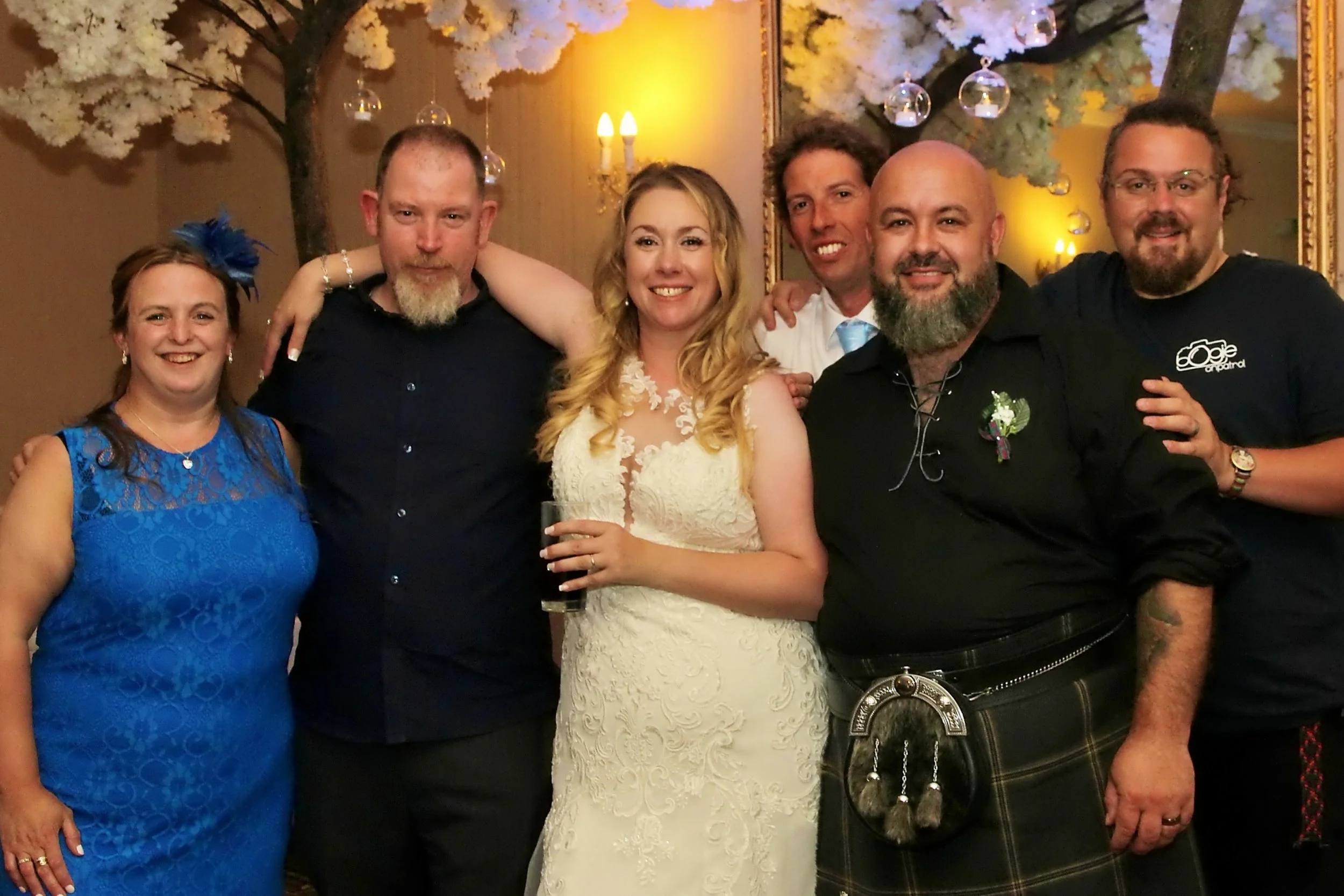 Group of seven people at a celebration, with a woman in a white lace dress holding a drink in the center, surrounded by friends in formal attire, in a decorated venue with white floral arrangements and hanging glass ornaments.