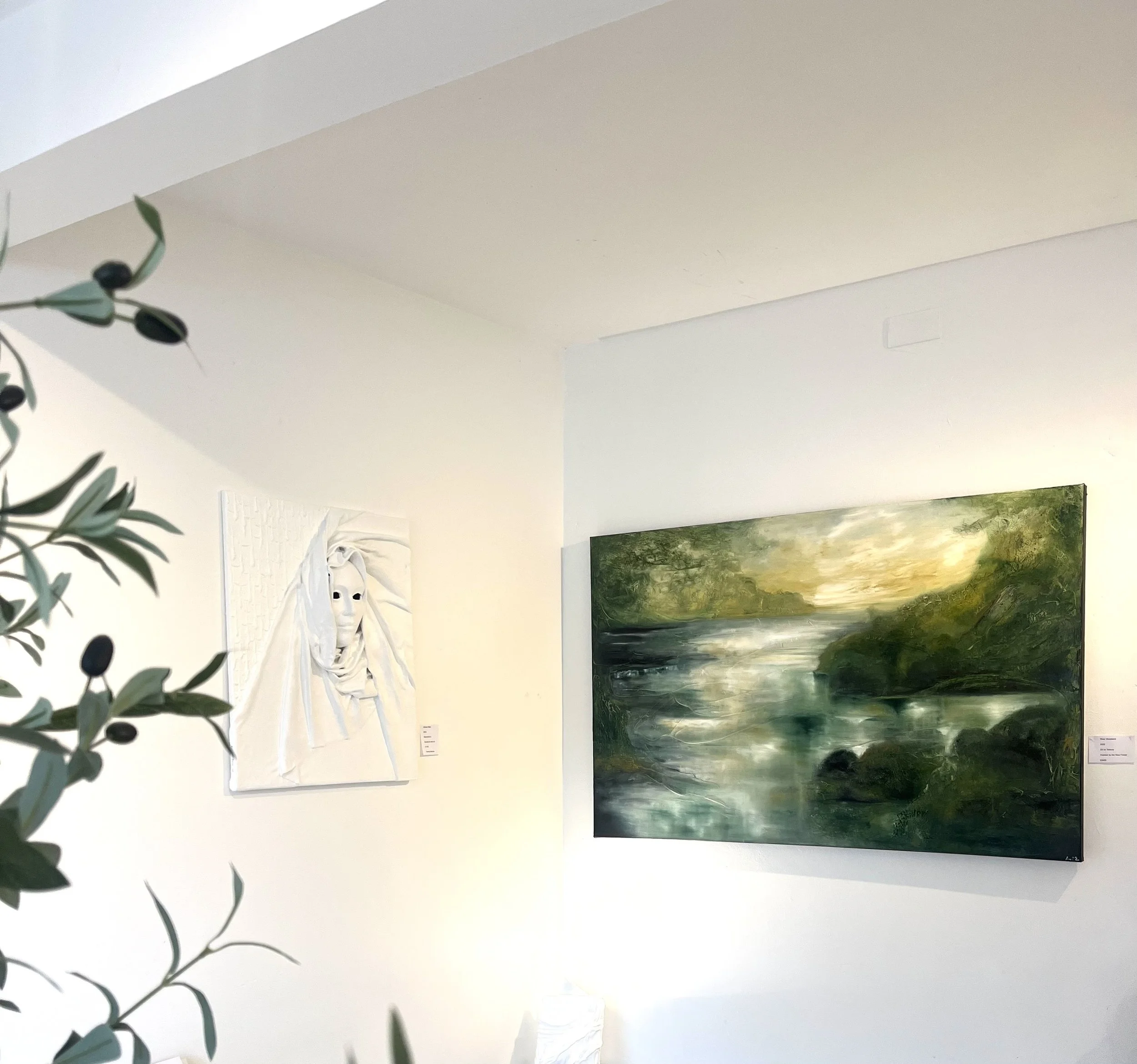 Art gallery with a landscape painting of a river and greenery on the right and a white sculpture of a face on the left wall, partially obscured by plant leaves in the foreground.