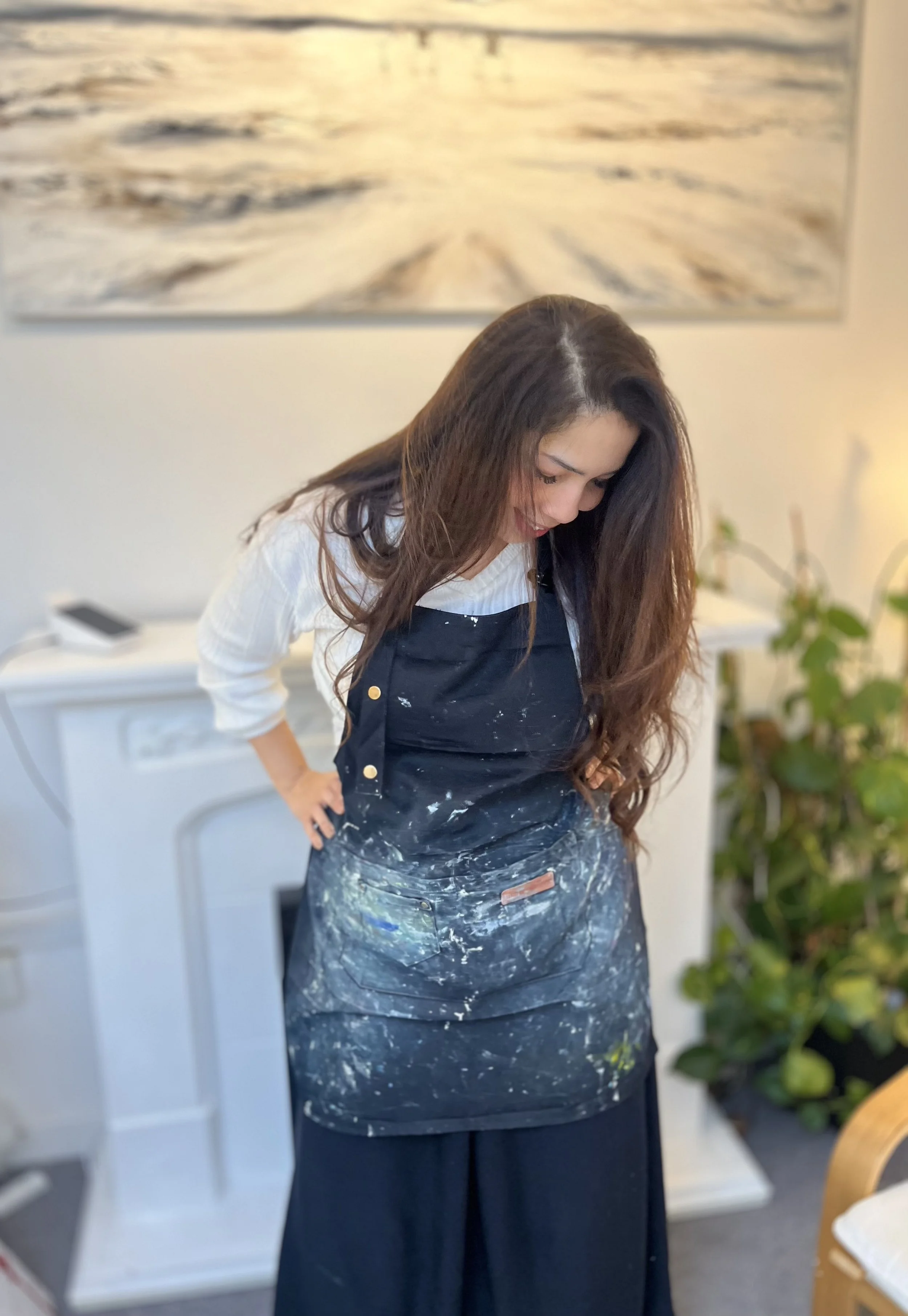 Woman with long brown hair looking down, wearing a white shirt and black apron splattered with paint or similar substance, in a room with a white dresser, a painting of a body of water, and green plants.