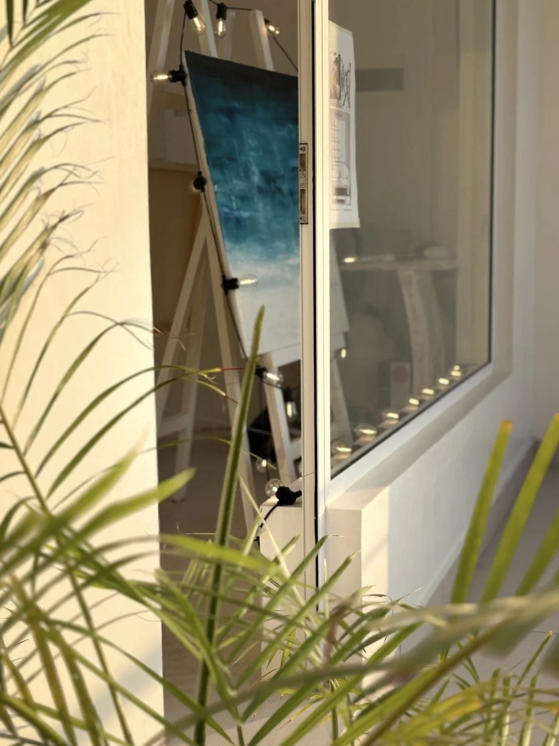 Close-up of green plant leaves in the foreground with a window reflecting a room with a piece of artwork on an easel inside.