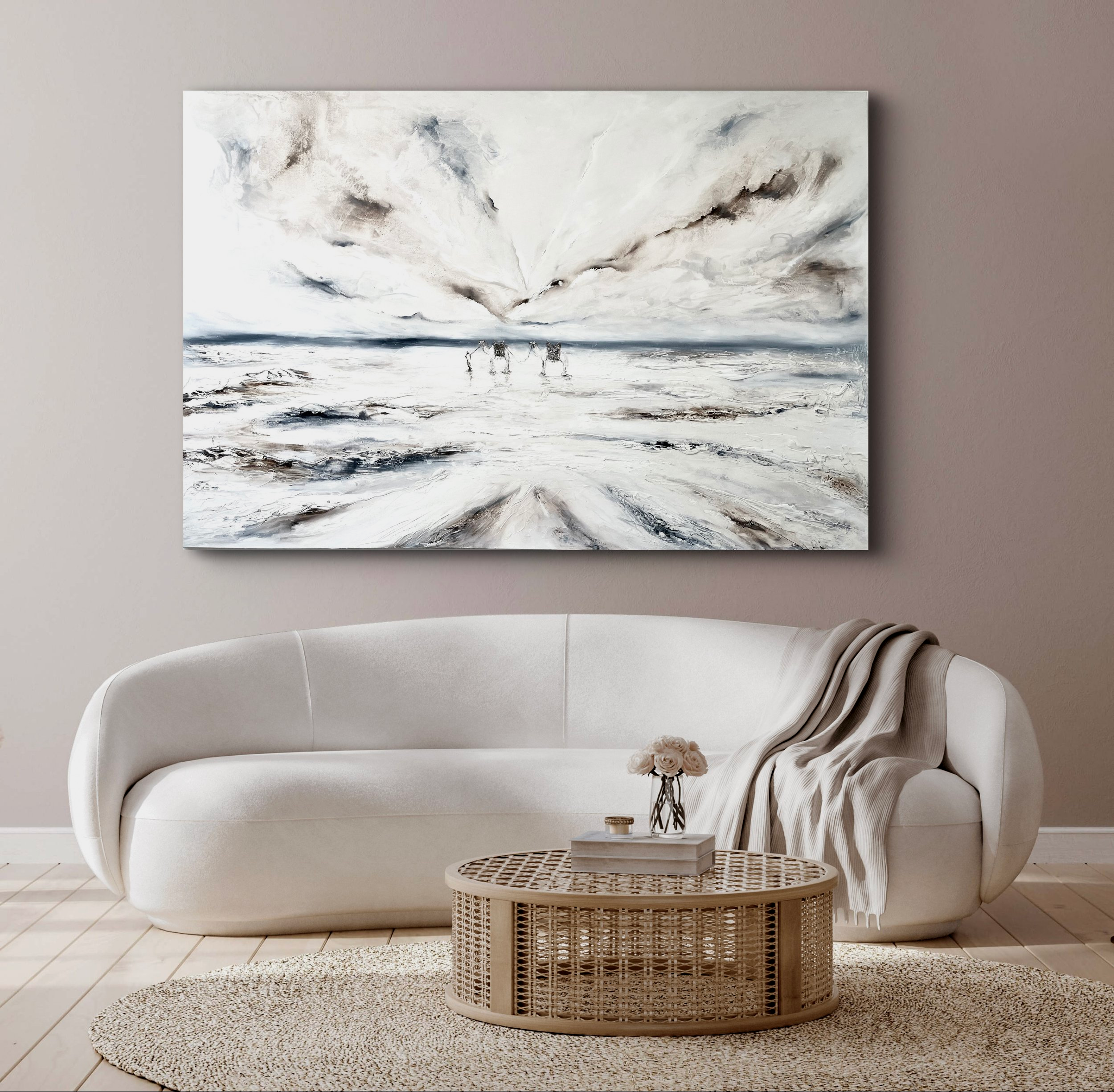 A modern living room with a white curved sofa, a round wicker coffee table with a vase of flowers, a beige throw blanket, a textured rug, and an abstract landscape painting of a snowy beach scene on the wall.