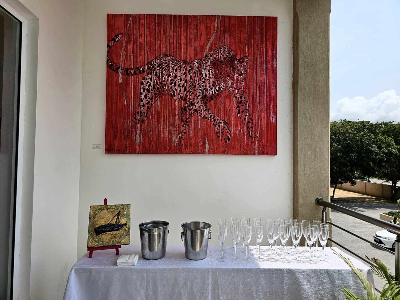 An art display with a large red canvas featuring a leopard walking, hanging on a white wall. A table below has two metal buckets, a small painting of a boat on a stand, and several empty champagne glasses.