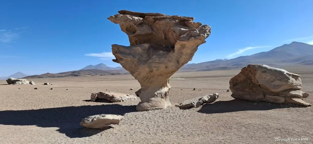 A desert landscape with large rock formations, including a prominent mushroom-shaped rock in the center, under a clear blue sky with distant mountains.