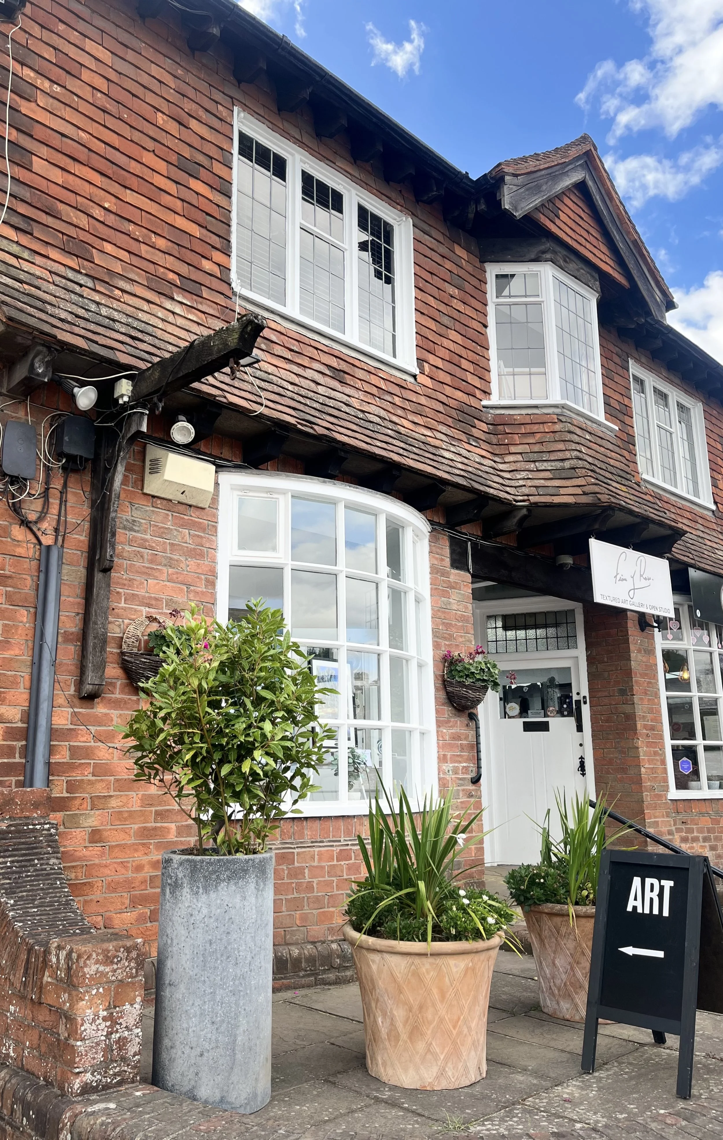 Brick art gallery building with large bay windows, potted plants outside, and a sign for art pointing left.