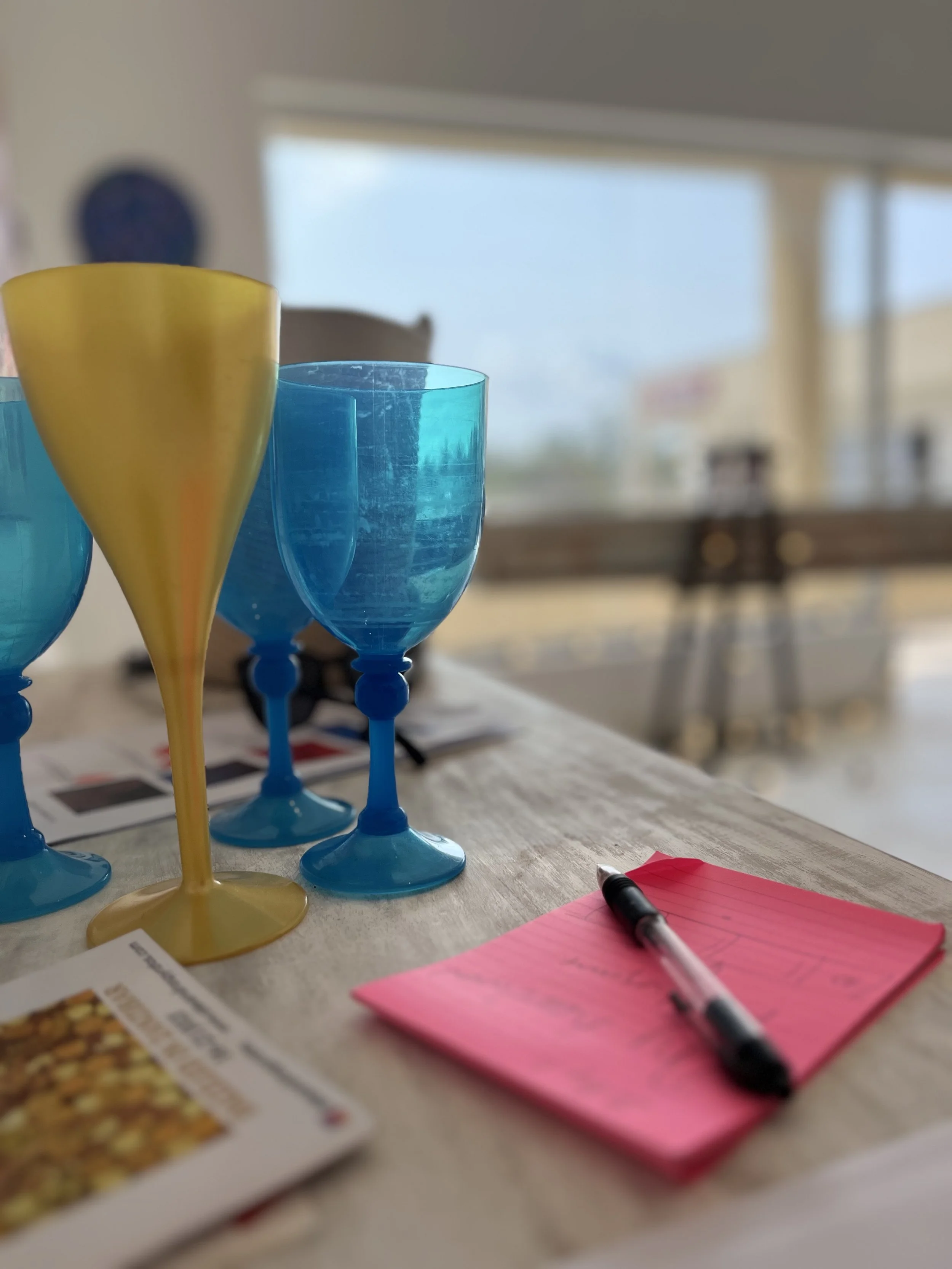 Colorful glasses, a pink notepad with a black pen, and a menu on a wooden table, with large windows and a blurred background.