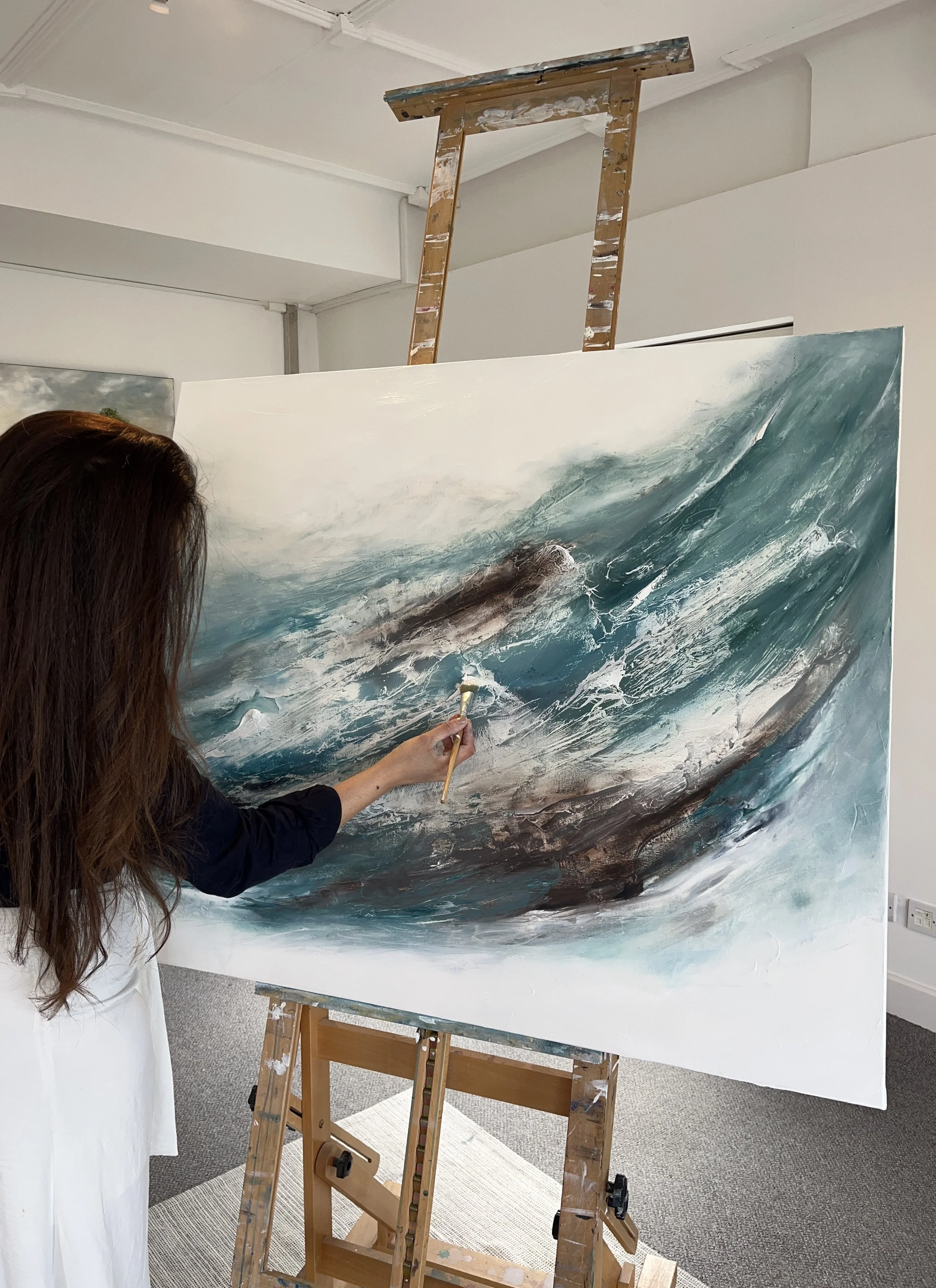 An artist painting a seascape of large ocean waves on a canvas in an indoor studio.
