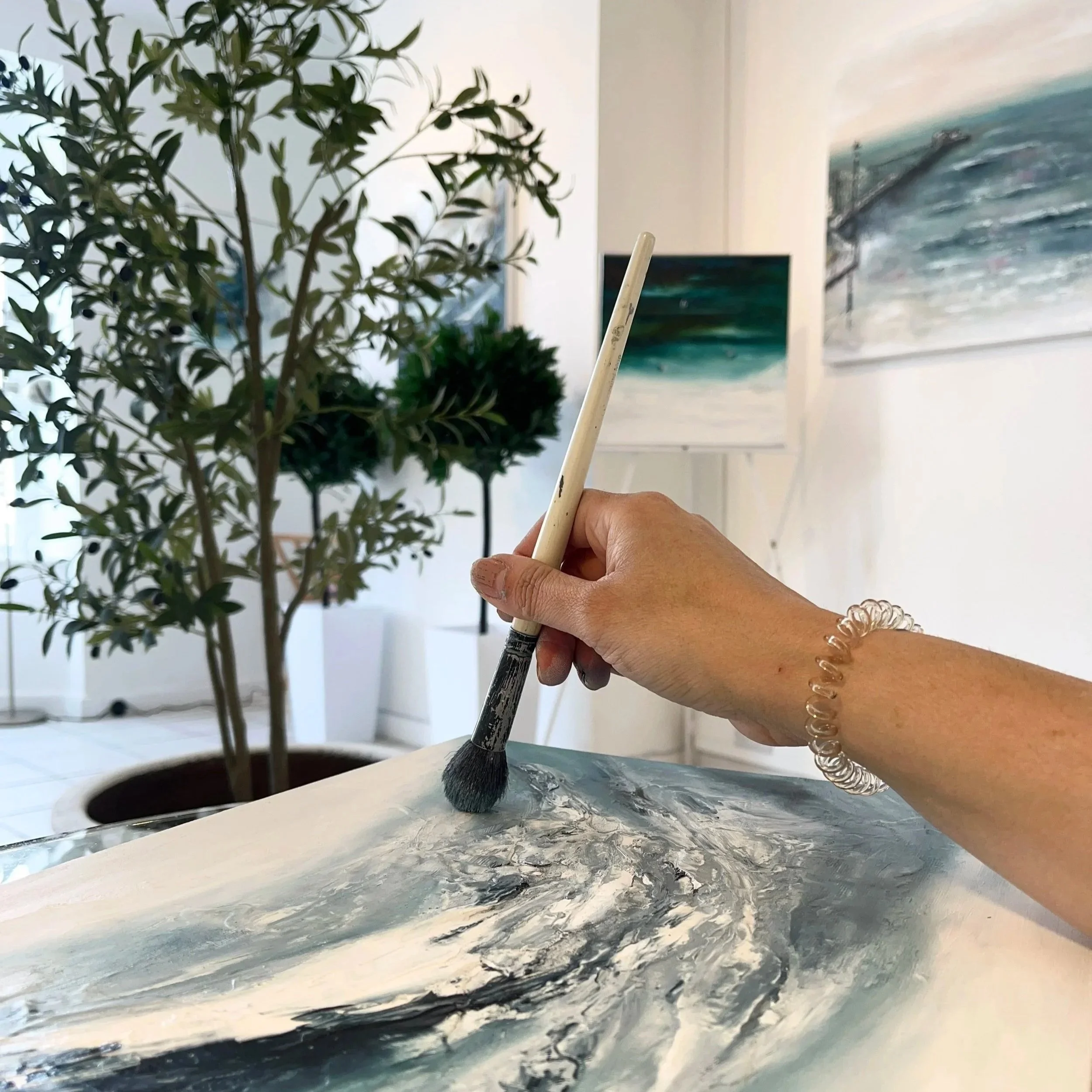 A woman at an art workshop and a paint-splattered apron stands smiling while painting on a canvas in a bright, indoor art studio. Other people are visible in the background, also working on art projects, with easels and art supplies around.