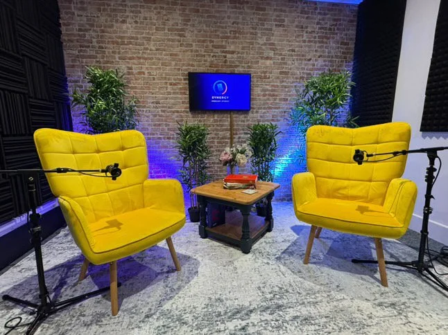 Two yellow armchairs with microphones, a wooden table with books and flowers, a television screen on a brick wall, and potted plants in a recording studio or podcast setup.