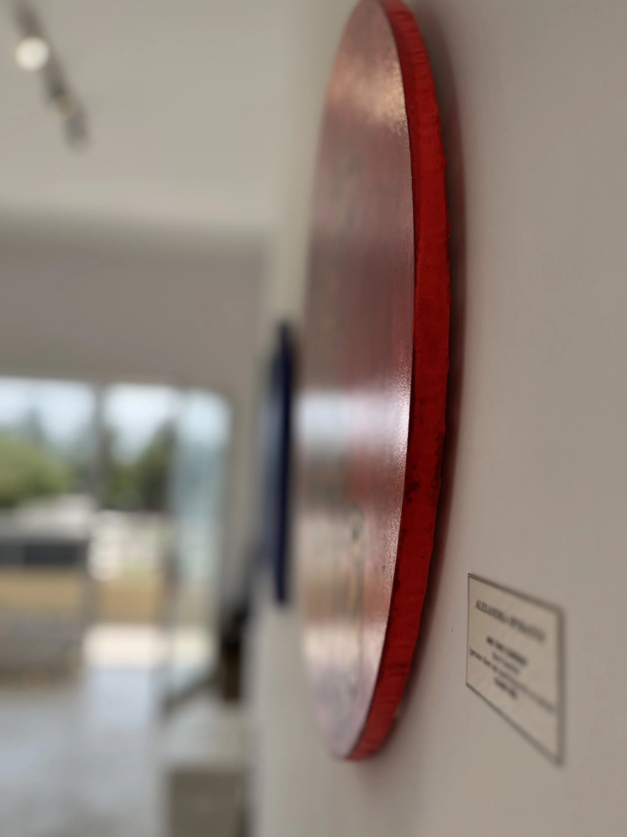 Close-up side view of a red circular artwork mounted on a white wall, with a blurred background.