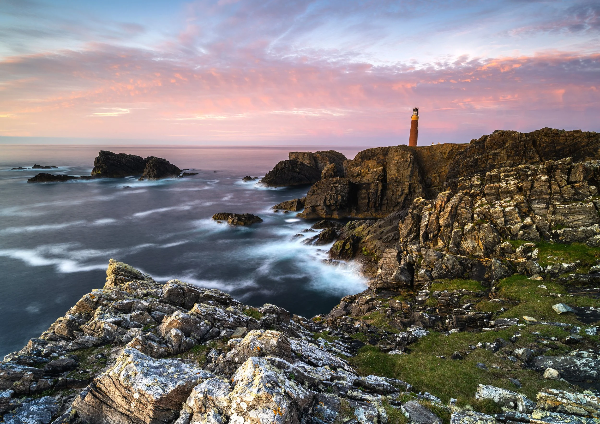 Butt of Lewis lighthouse above rugged cliffs with soft pink light and long‑exposure sea motion.