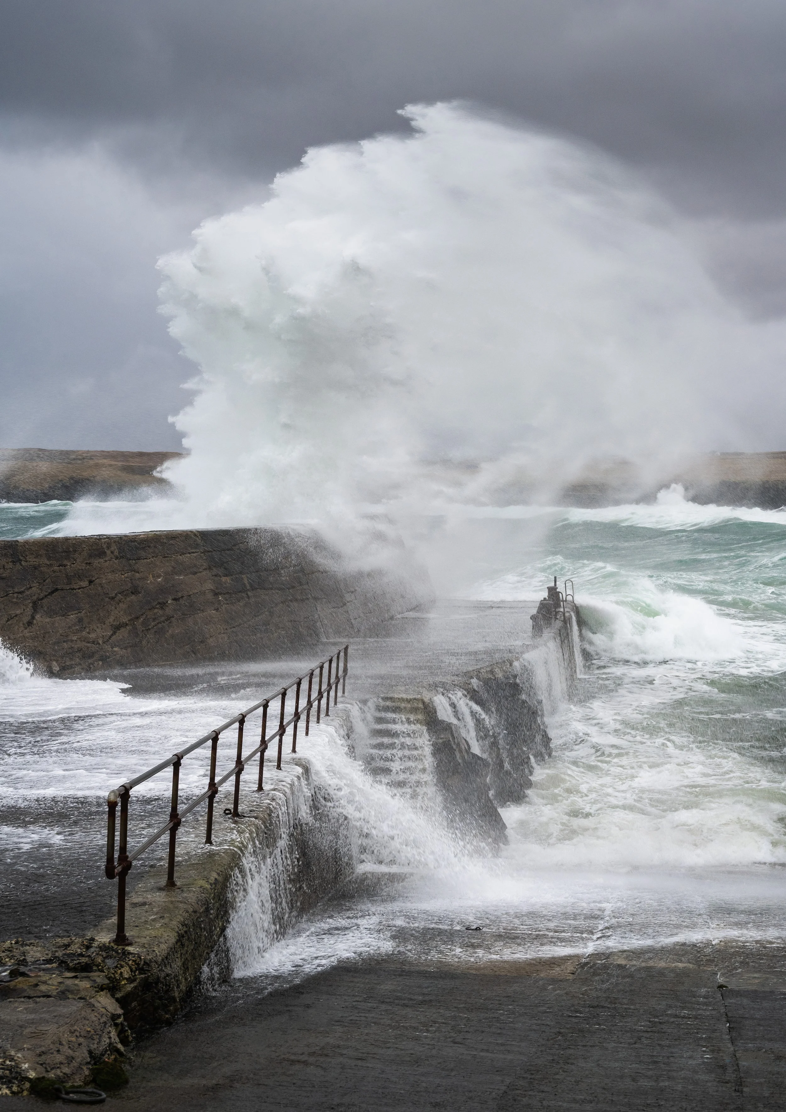 Port Of Ness, Isle of Lewis. Waves crashing against a rocky shoreline during a storm, with a pier and railing in the foreground.