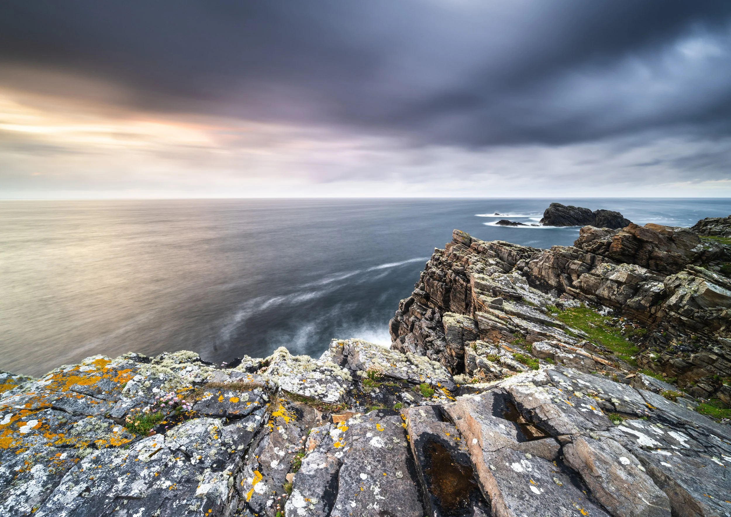 Rocky cliff overlooking the ocean with dark stormy clouds and some light near the horizon.