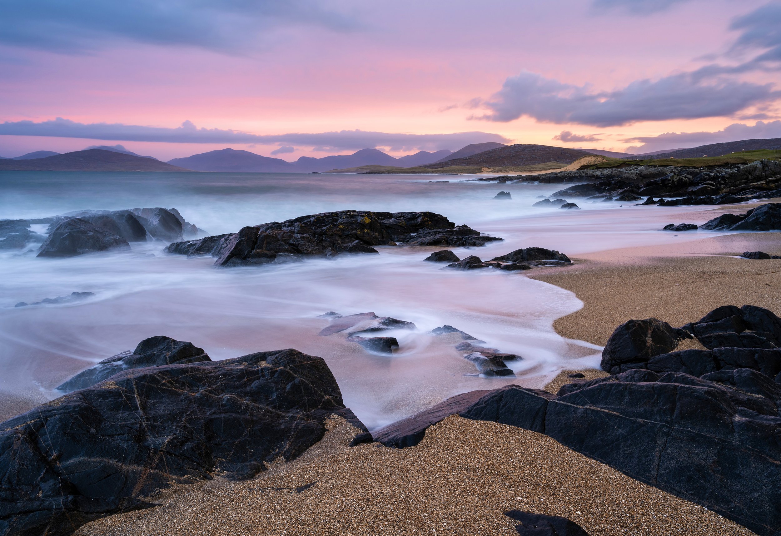 Small beach, isle of Harris  during sunrise with pink and purple sky, rocky shoreline, and gentle waves washing over the sand.