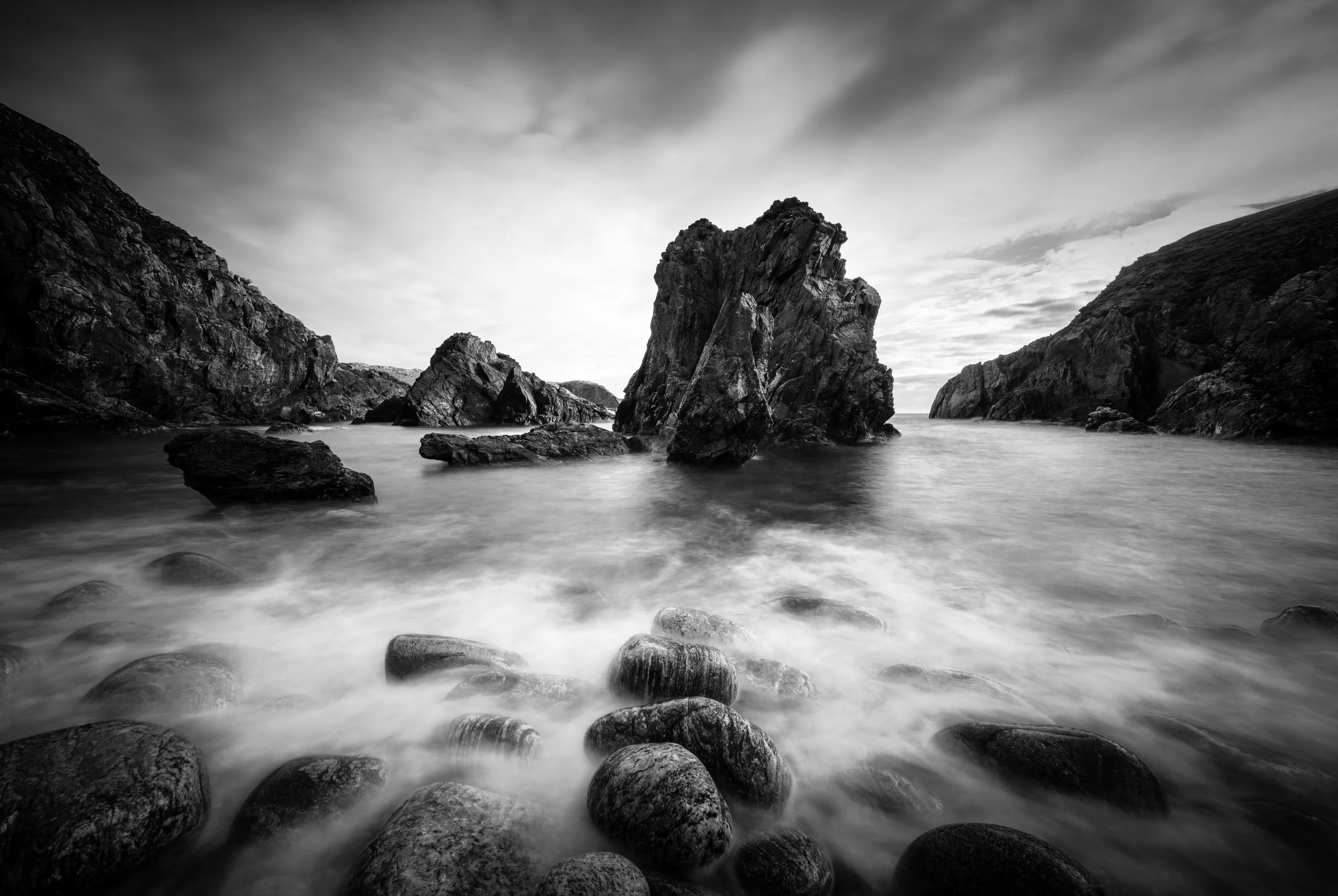 Long‑exposure black‑and‑white seascape from the Outer Hebrides, with rounded foreground rocks, jagged sea stacks and soft Atlantic movement