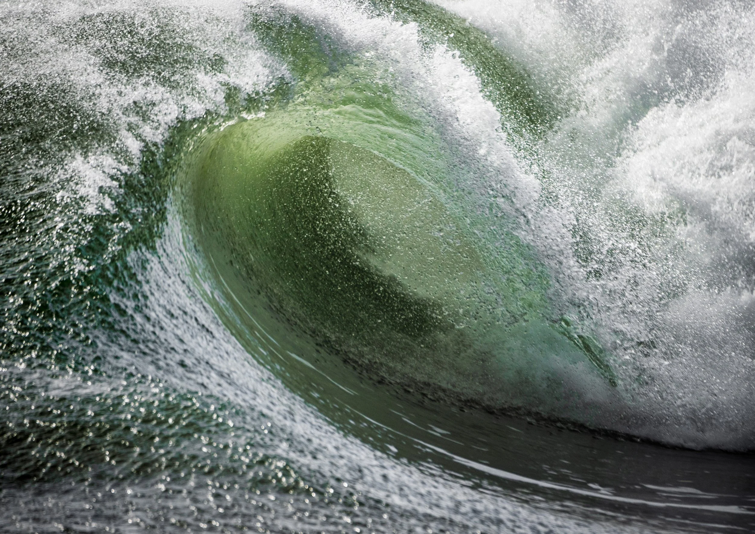 Green wave breaking in close‑up, with curved water, textured spray and soft light highlighting the movement of the sea.