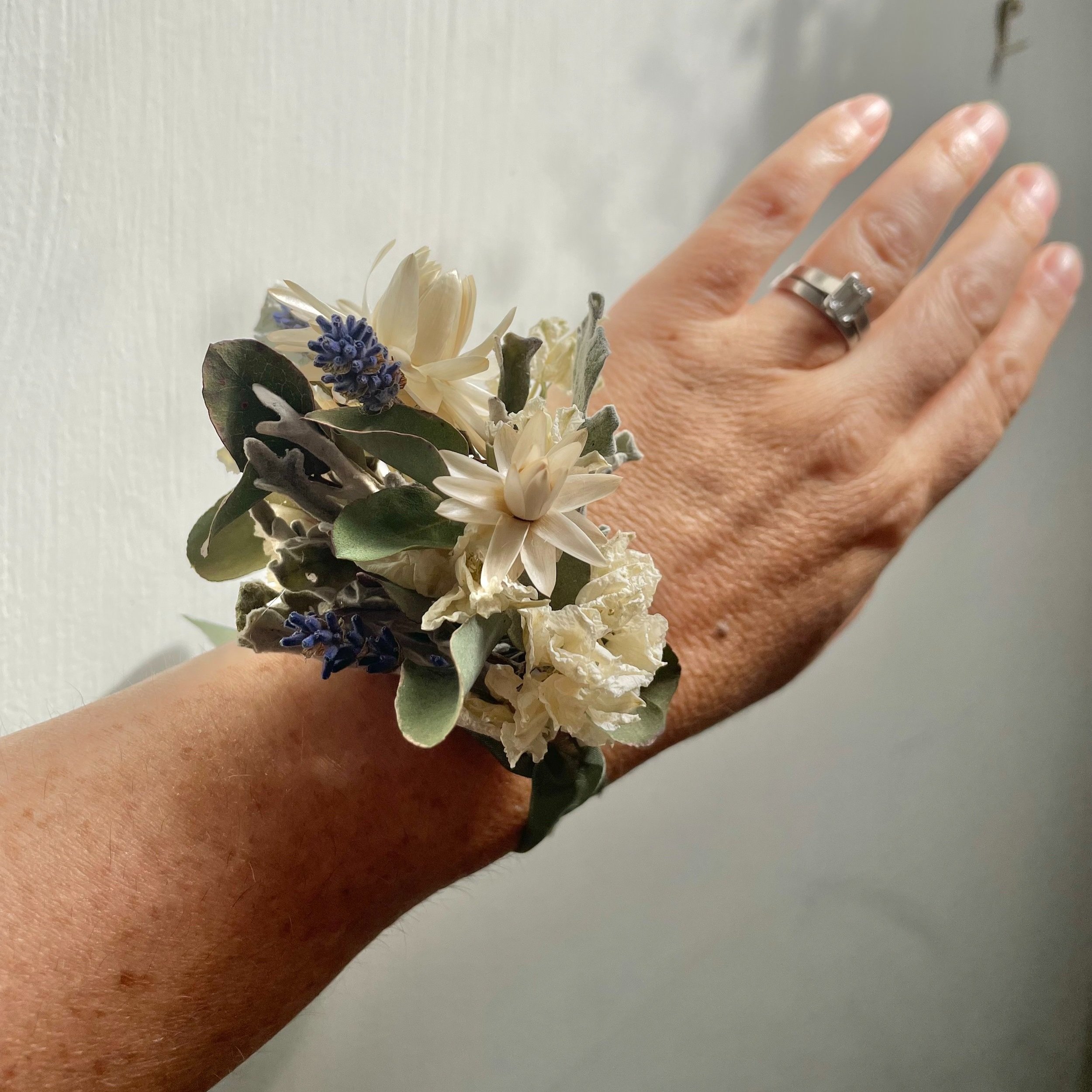 A dried flower corsage worn on a wrist. Grey green foliage, ivory strawflowers and sprigs of lavender are arranged and tied with a sage green ribbon.