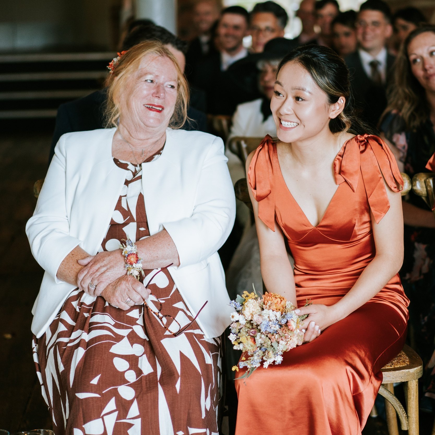 two female members of the bridal party sit together, one wears a dried flower wrist corsage and the other, a bridesmaid in a brunt orange silk dress, has her dried flower posy in her lap.