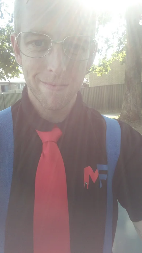 A man wearing glasses, a black shirt with a logo, a red tie, and a blue backpack taking a selfie outdoors in bright sunlight with trees, a fence, and buildings in the background.