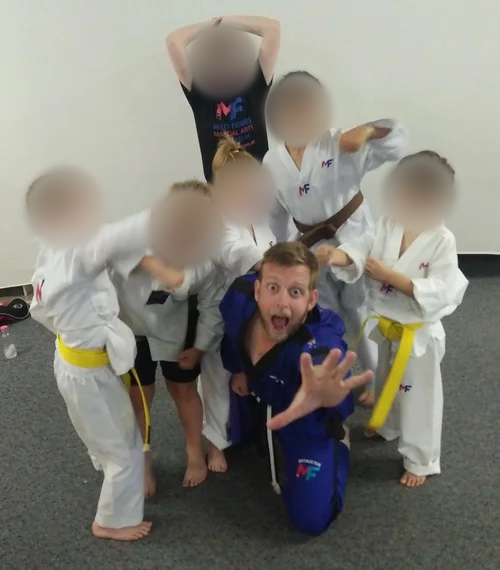 Group of children and instructor in martial arts uniforms posing playfully in a dojo. Children’s faces blurred for privacy.